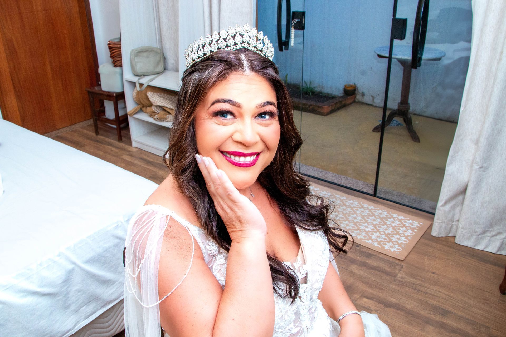 Noiva de vestido branco e tiara sorri, com a mão na bochecha, perto da porta de vidro.