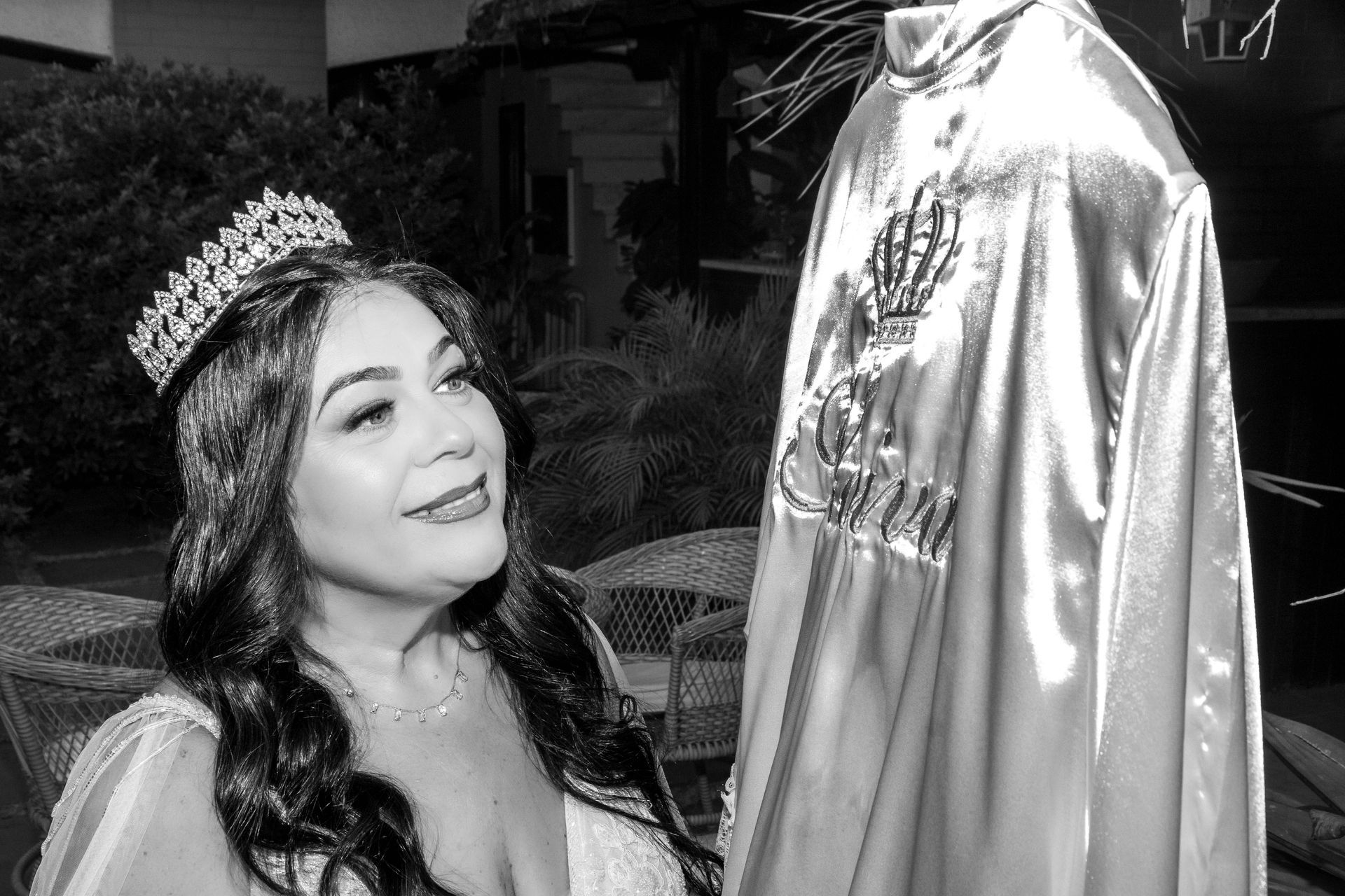 Mulher com tiara sorri para uma capa de cetim com um emblema de coroa pendurado nas proximidades, em ambiente externo.