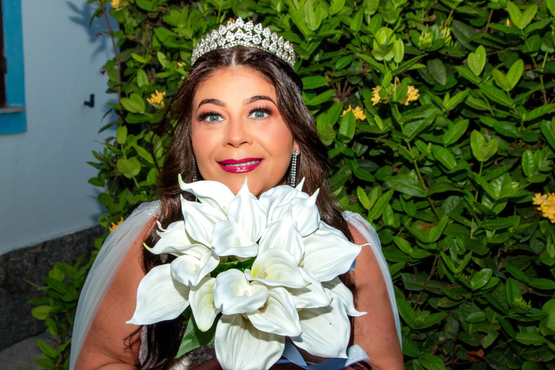 Noiva com tiara segura um buquê de flores brancas, sorrindo ao ar livre perto de folhagens verdes.