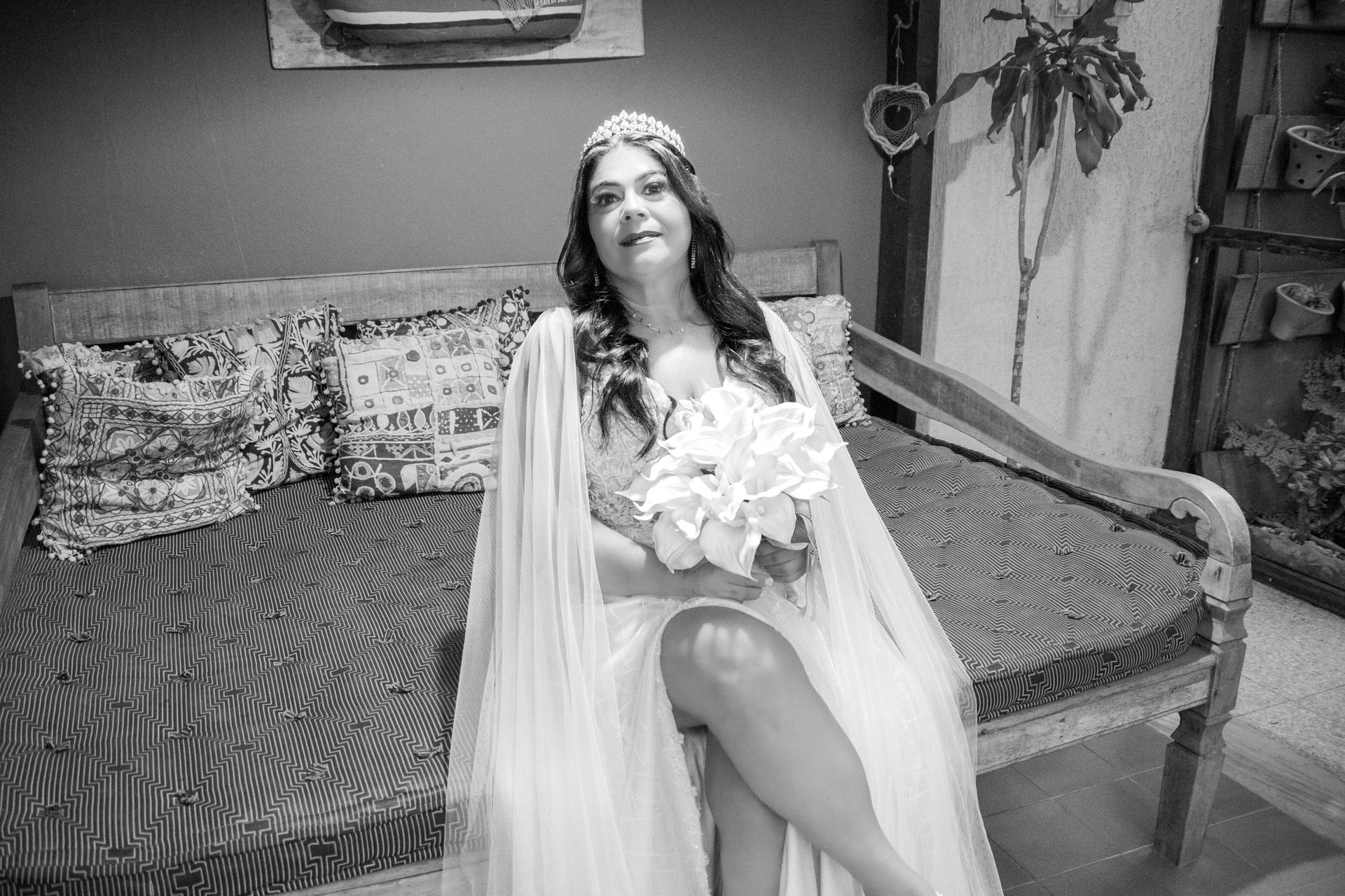 Mulher vestida de noiva segurando flores, sentada em um sofá, usando uma coroa e capa.