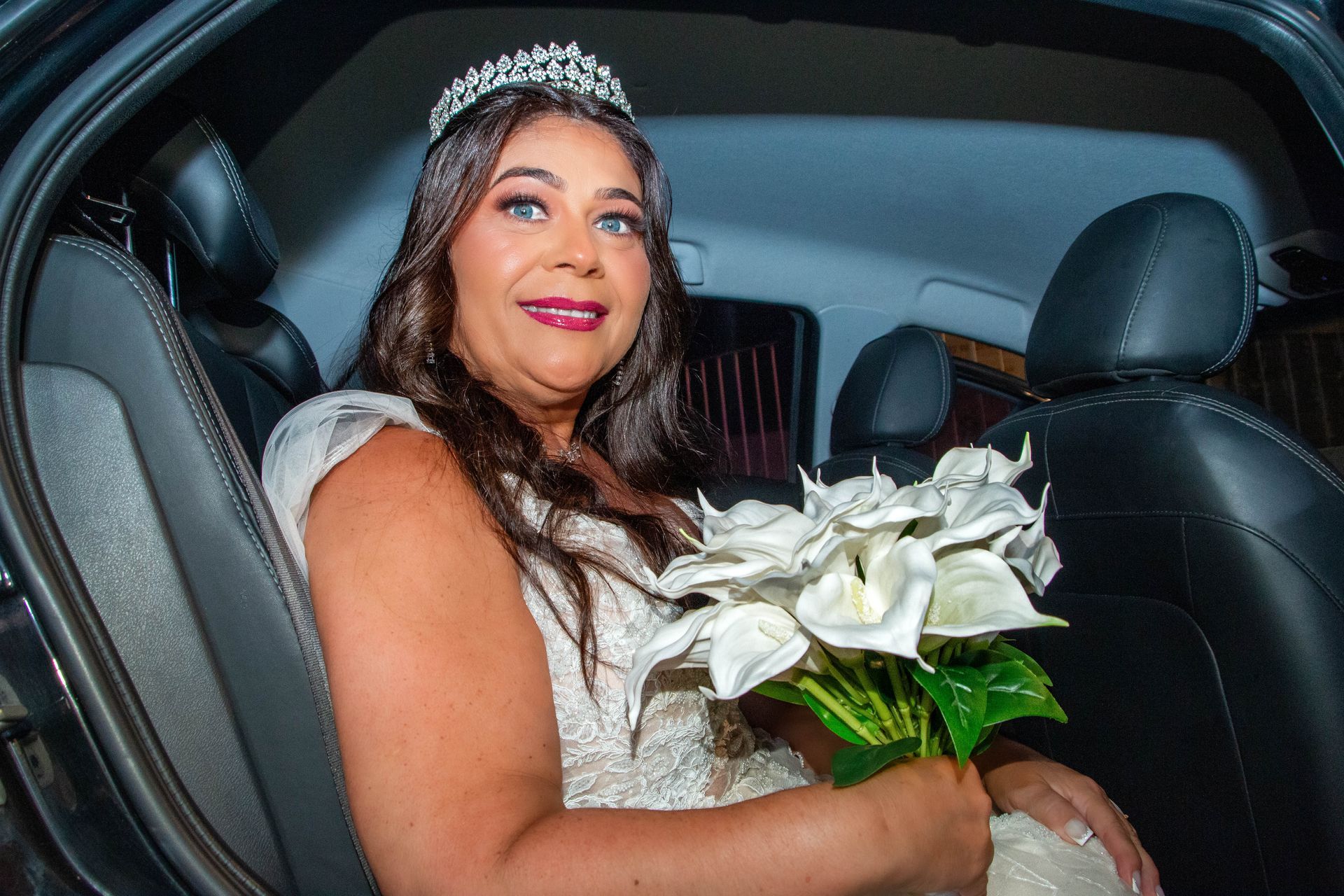 Noiva no carro segurando flores brancas, usando uma tiara e sorrindo.
