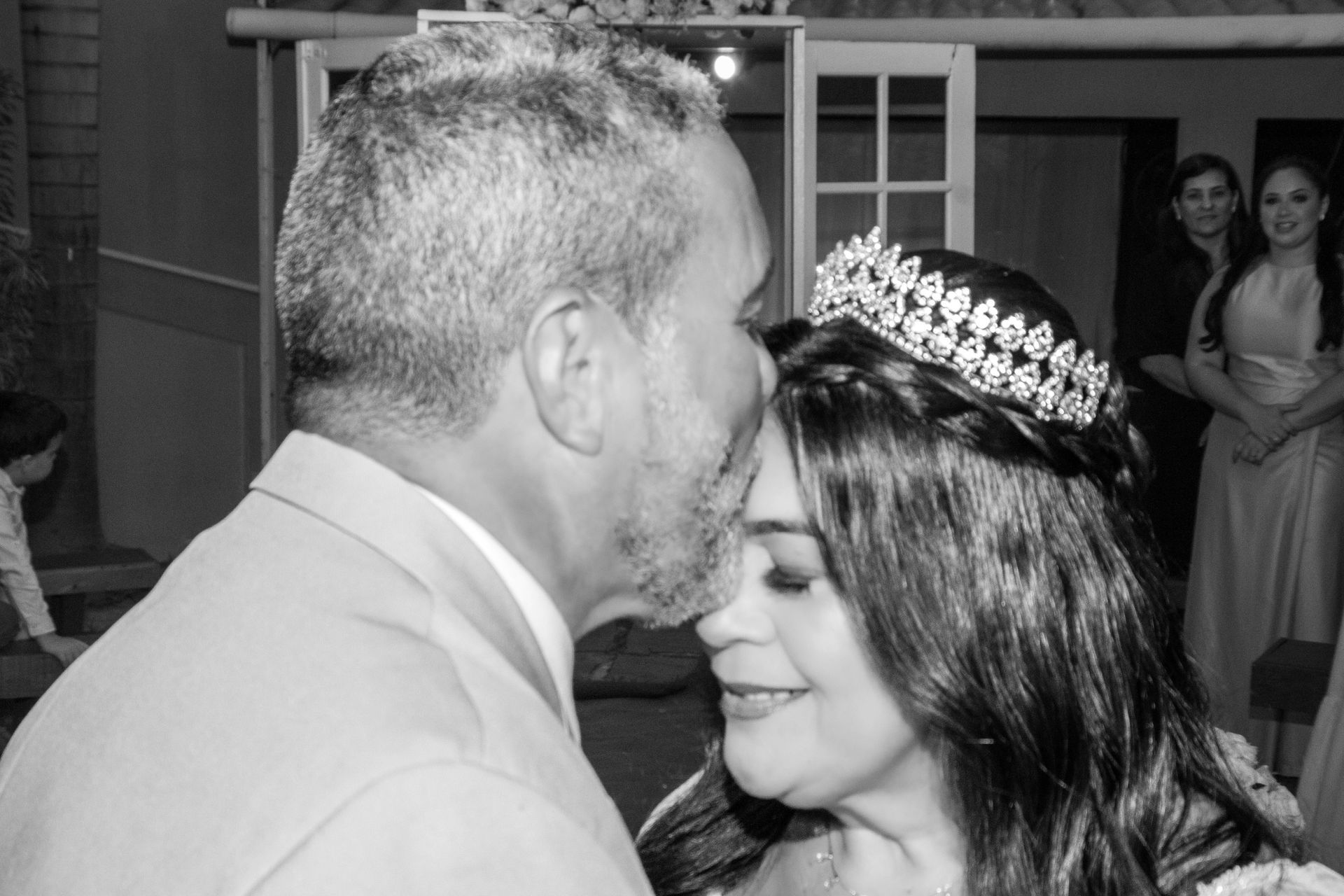 Homem beijando a testa de uma mulher; ela usa uma tiara. Outros observam ao fundo.