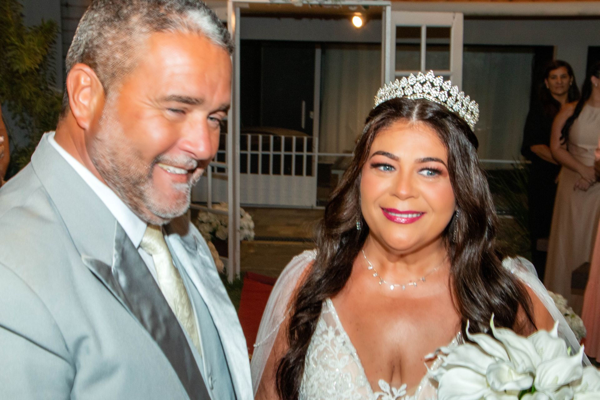Noivos em casamento. A noiva usa tiara e vestido; o noivo, terno. Eles sorriem do lado de fora de um prédio.