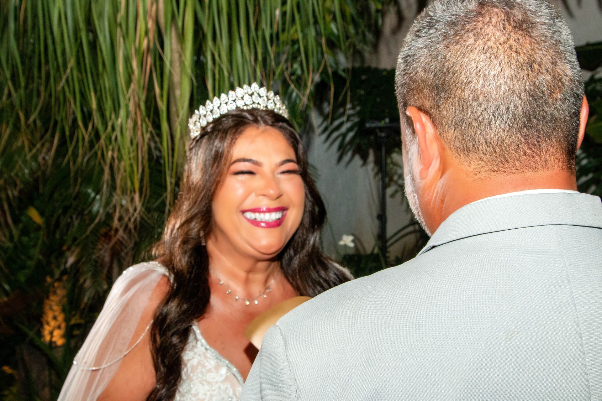 Noiva com tiara sorri para uma pessoa, provavelmente o noivo, em um jardim.