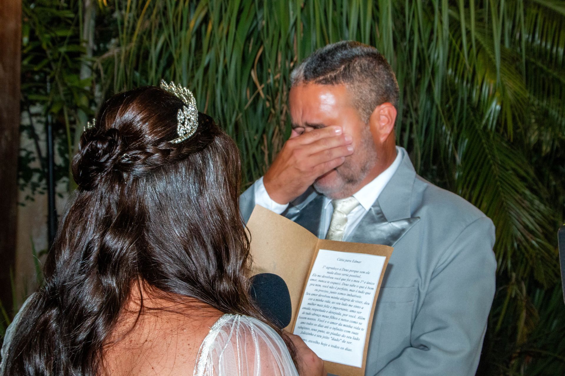 Noivo de terno cinza enxugando lágrimas, lendo os votos. Noiva de cabelos longos, lendo junto. Ambiente externo, noite.