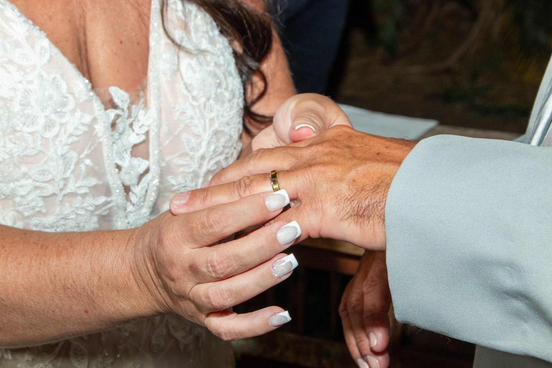 Mulher colocando aliança no dedo do homem durante uma cerimônia de casamento. Ela usa um vestido de renda branca. Ele usa um terno cinza.