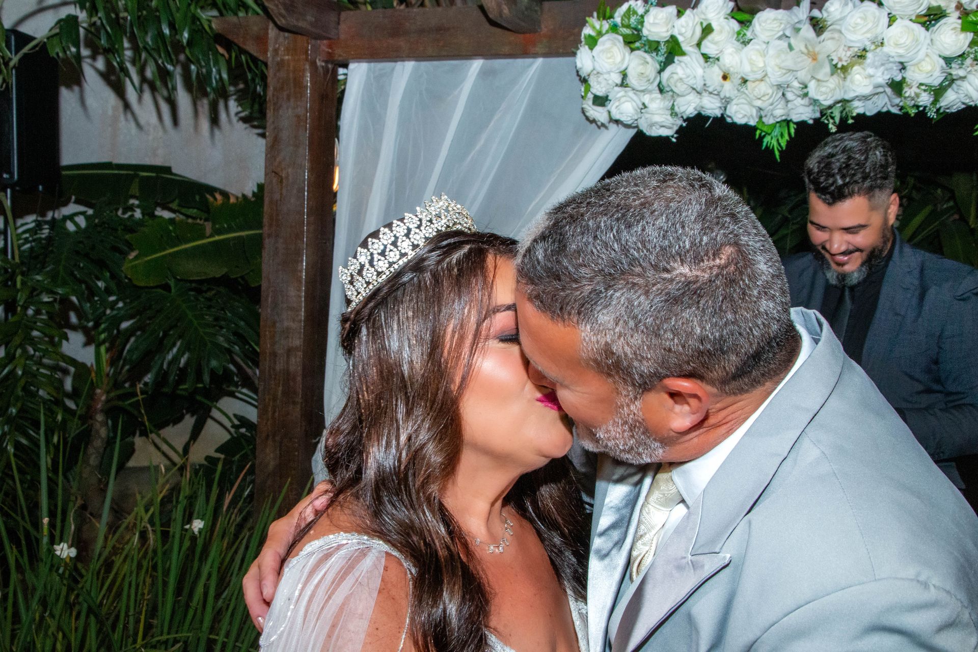 Casal se beijando sob um arco floral, homem de terno sorri ao fundo.