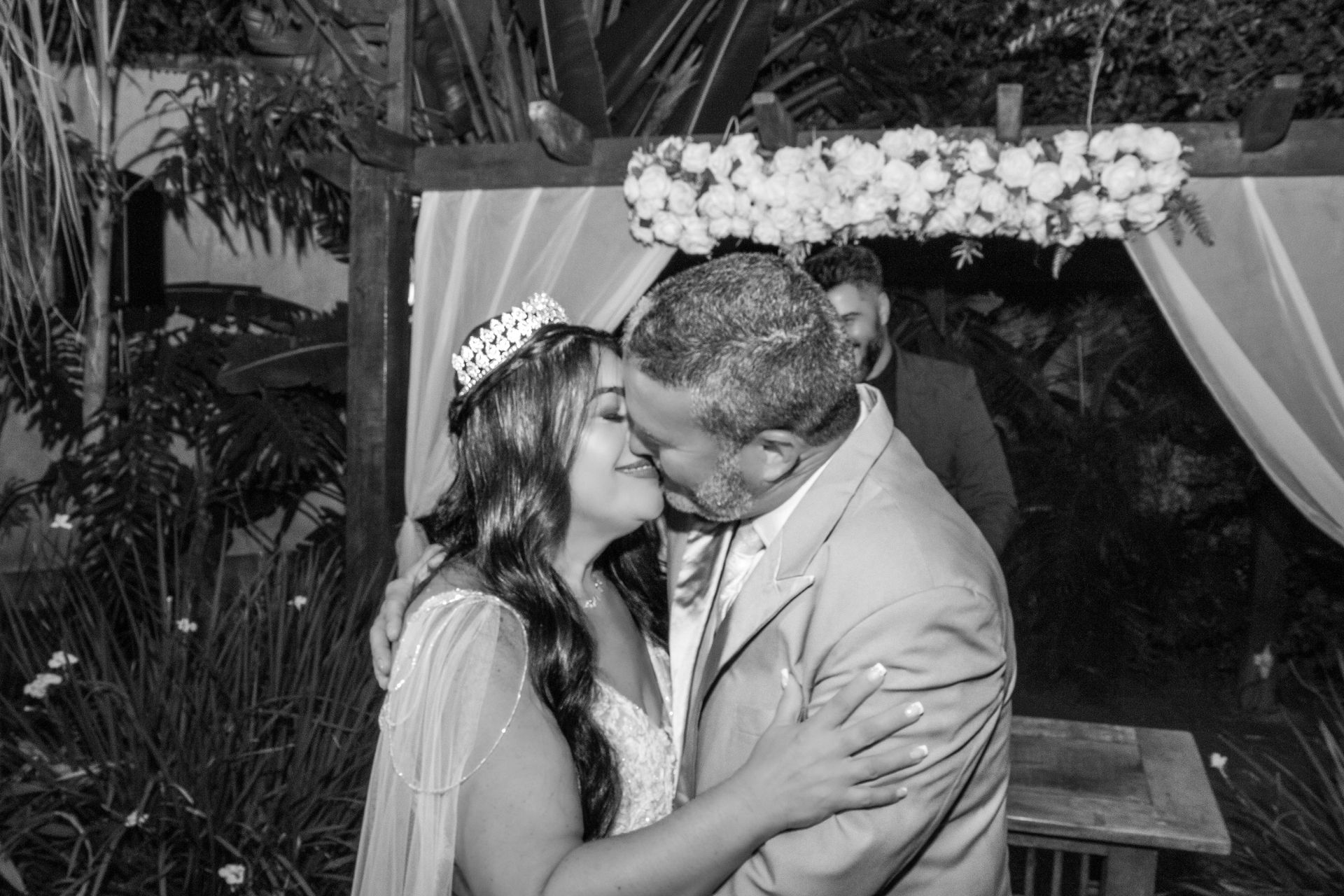 Noivos se beijando no altar, arco floral, ambiente externo, preto e branco.