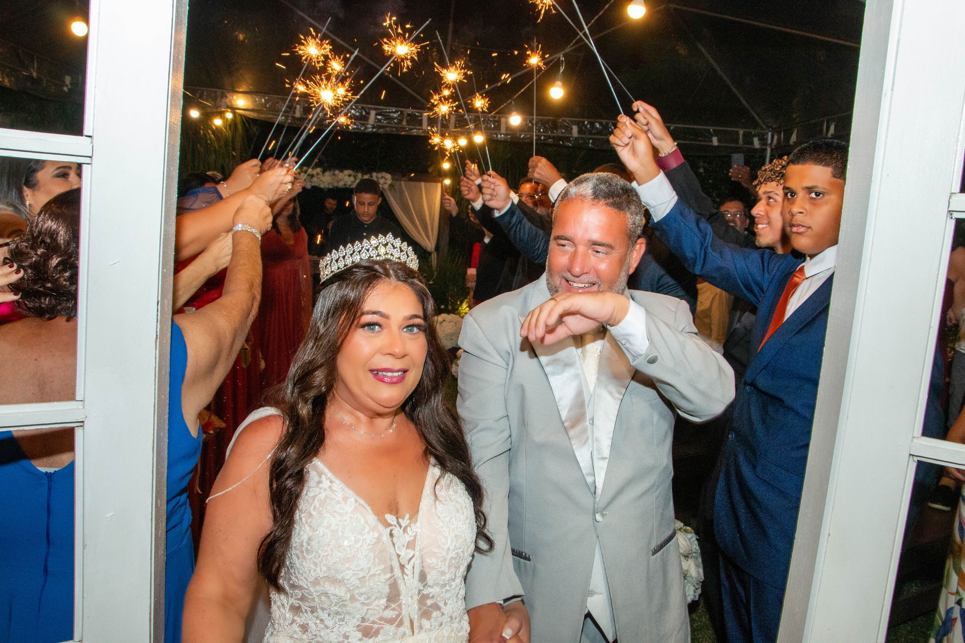 Recém-casados caminhando por uma saída de fogos de artifício; convidados seguram fogos de artifício no alto, em um jardim.