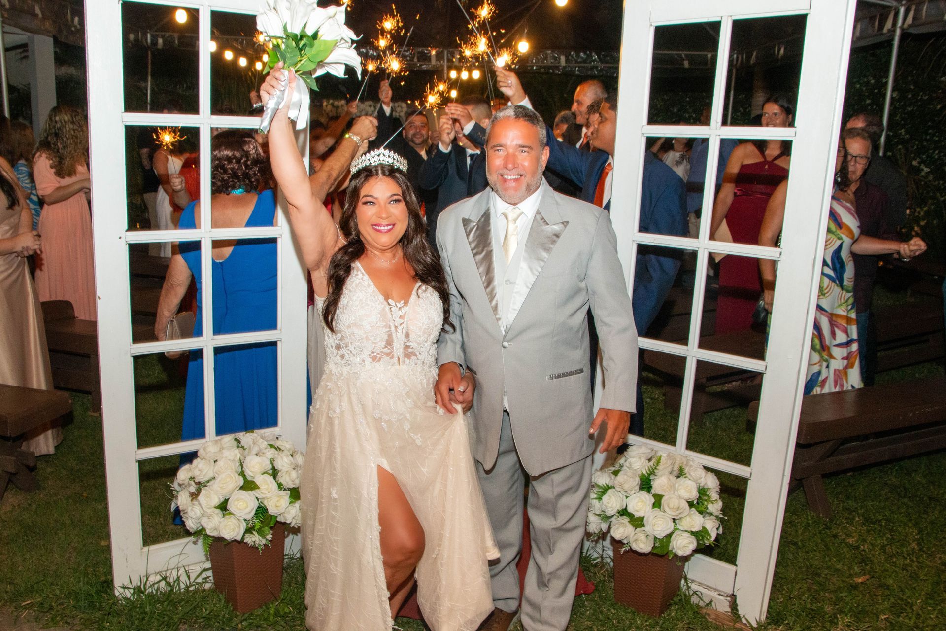 Noiva e noivo saindo da cerimônia de casamento através de uma moldura branca. Noiva ergue o buquê, noivo sorri. Ambiente externo.