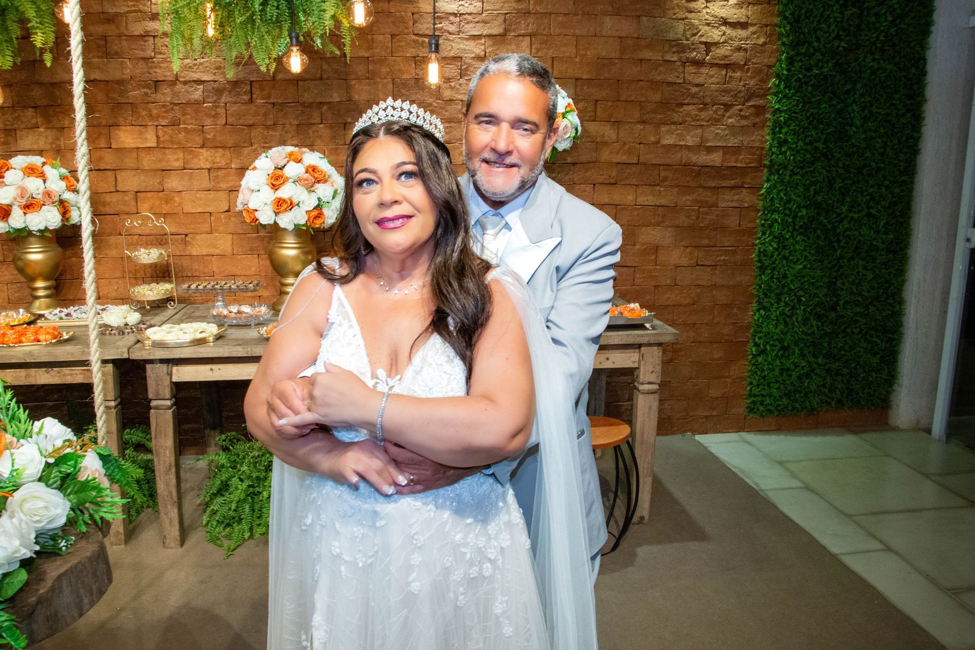 Casal se abraçando em uma recepção de casamento; a noiva usa um vestido branco e tiara, o noivo usa um terno claro.