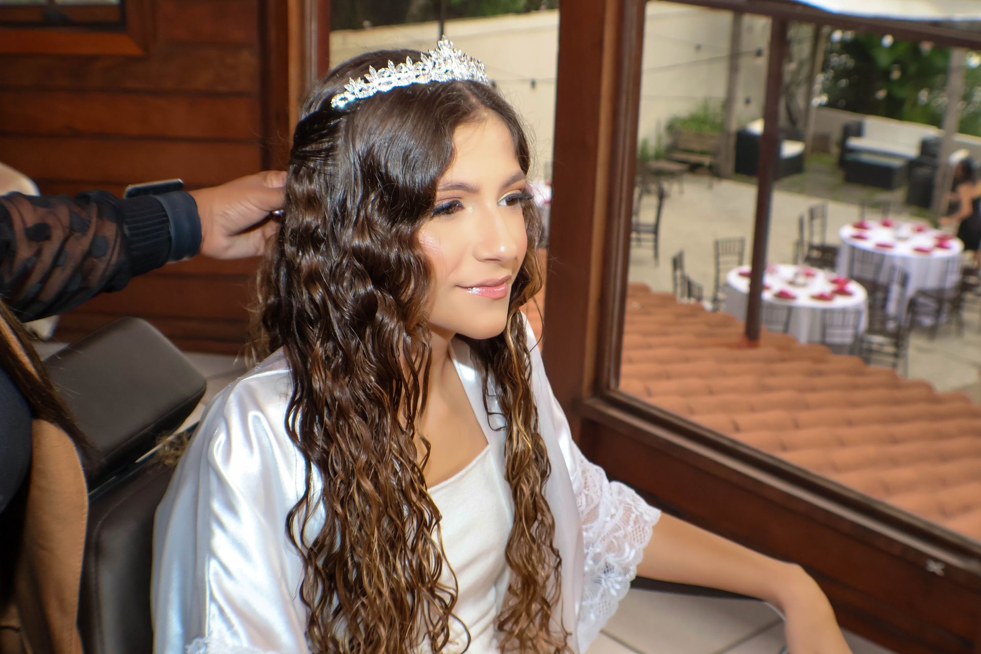 Mulher com cabelo castanho cacheado usando uma tiara e um robe branco; pessoa ajeitando o cabelo; evento ao ar livre ao fundo.