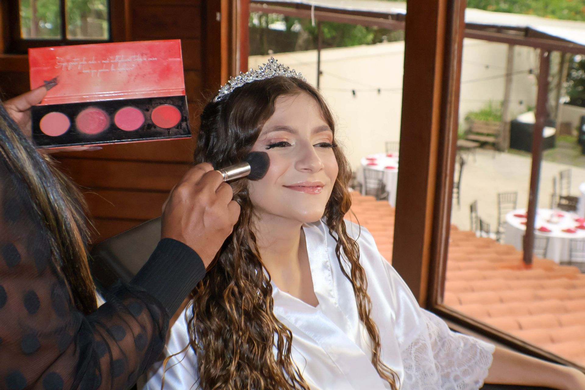 Mulher maquiada com paleta de blush. Ambiente externo com mesas.