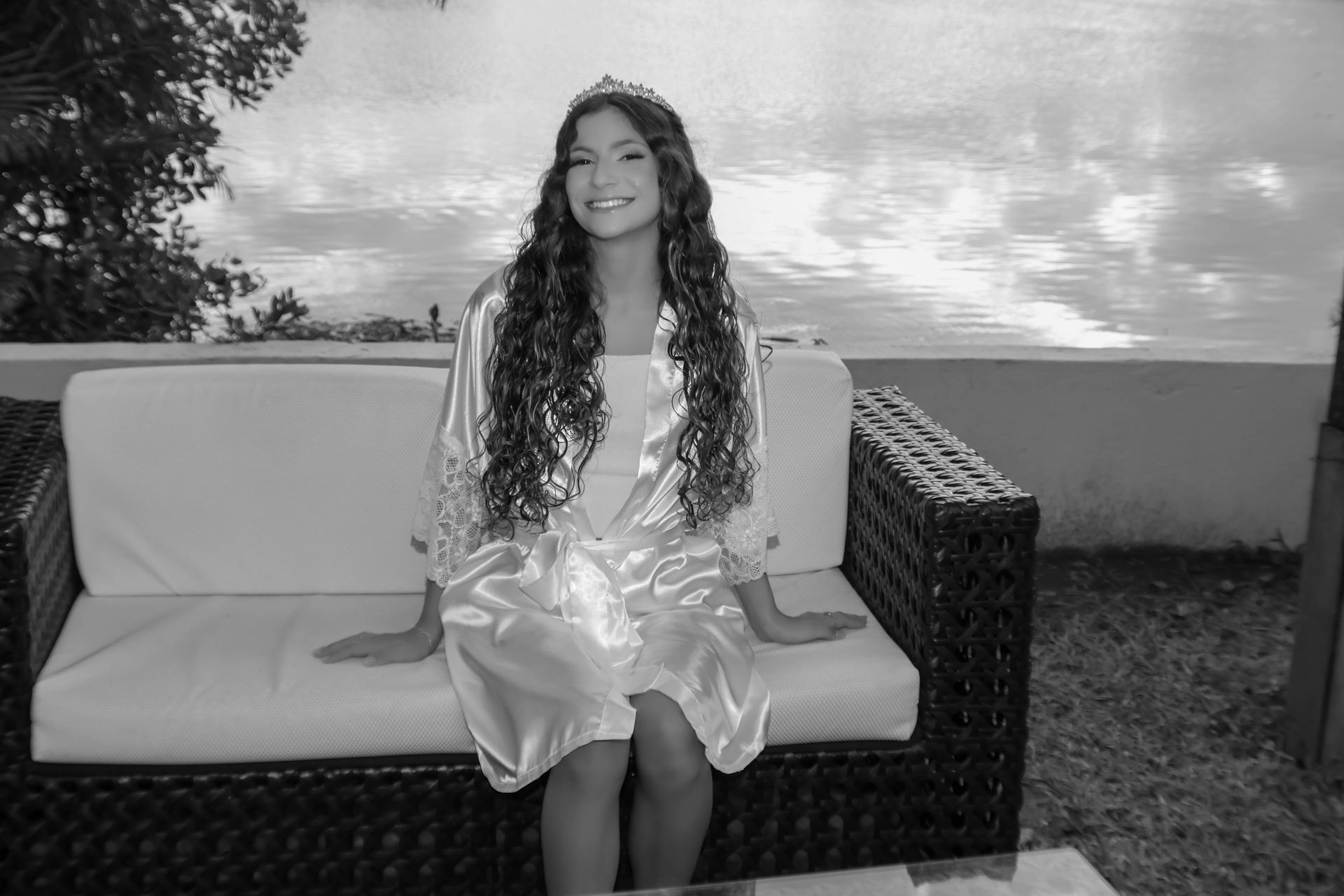 Mulher de roupão e tiara sentada em um sofá de vime, sorrindo. Cenário externo com água e céu ao fundo.