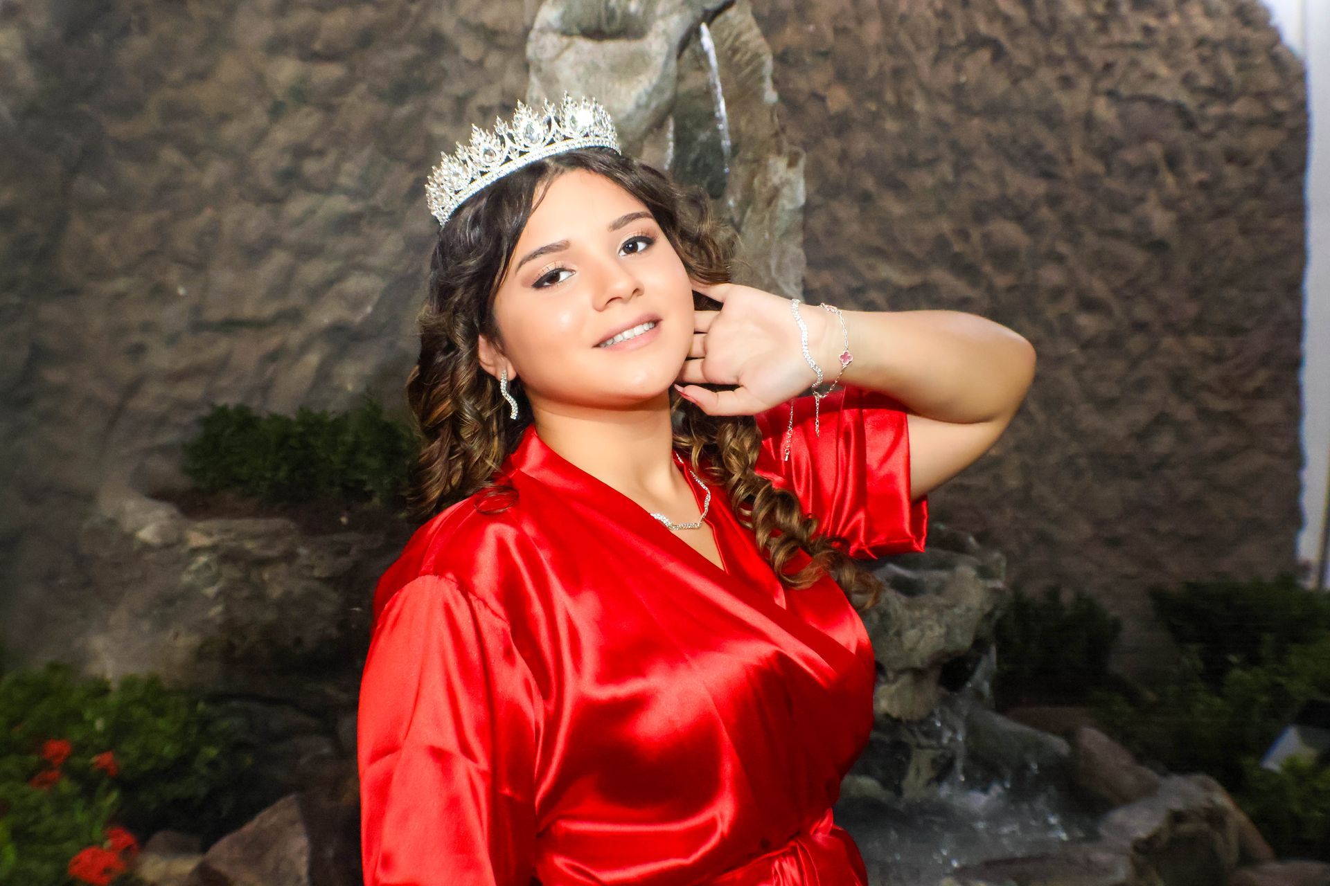 Pessoa em túnica de cetim vermelho usando uma tiara, sorrindo, perto de uma pequena cachoeira.