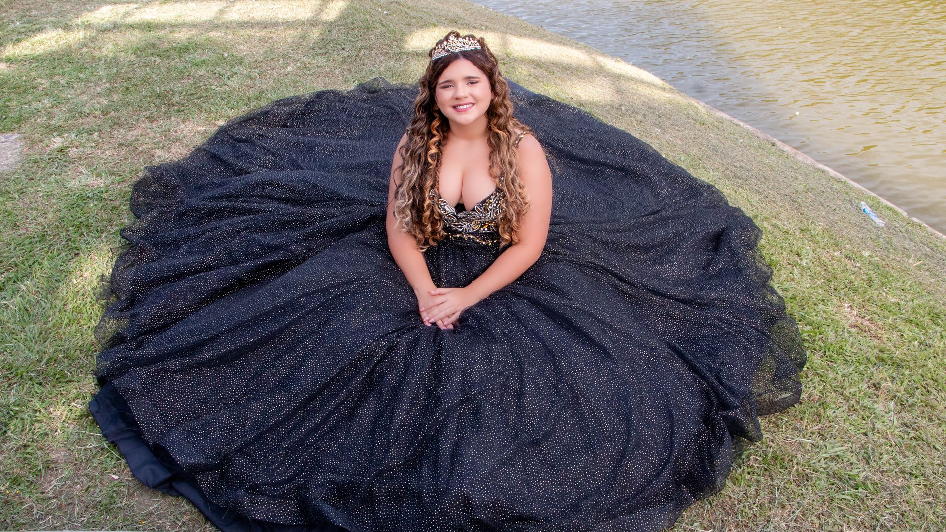 Mulher com um vestido de baile preto brilhante e coroa sentada na grama.