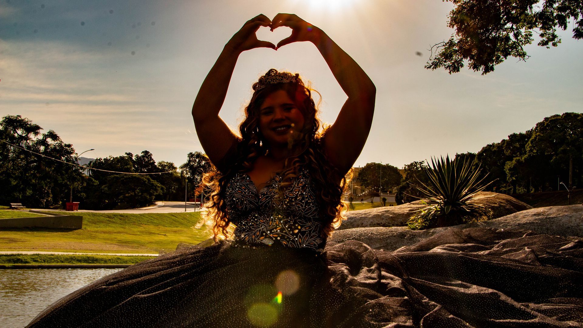 Mulher formando um coração com as mãos, silhuetada pelo sol, usando um vestido e uma coroa.