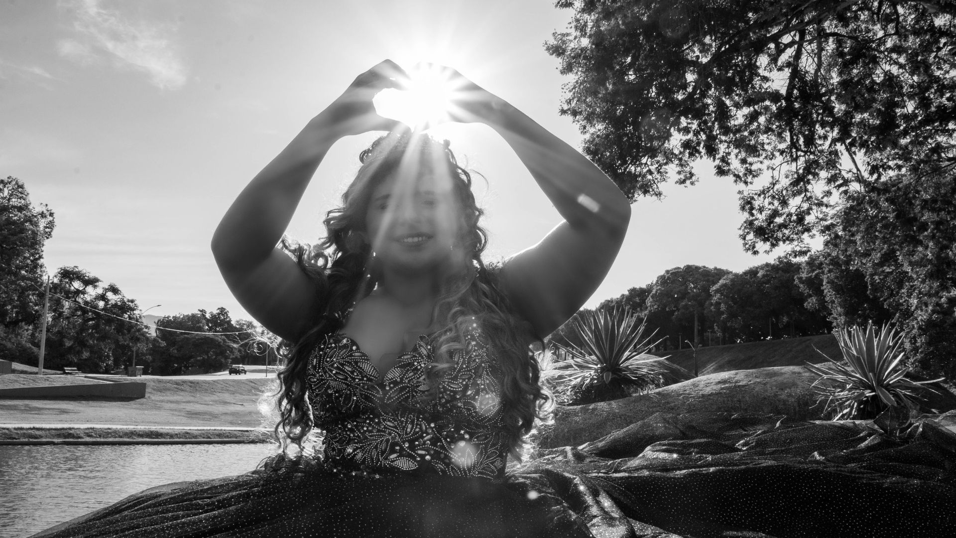 Pessoa fazendo um formato de coração com as mãos ao redor do sol, em pé perto da água e das árvores.