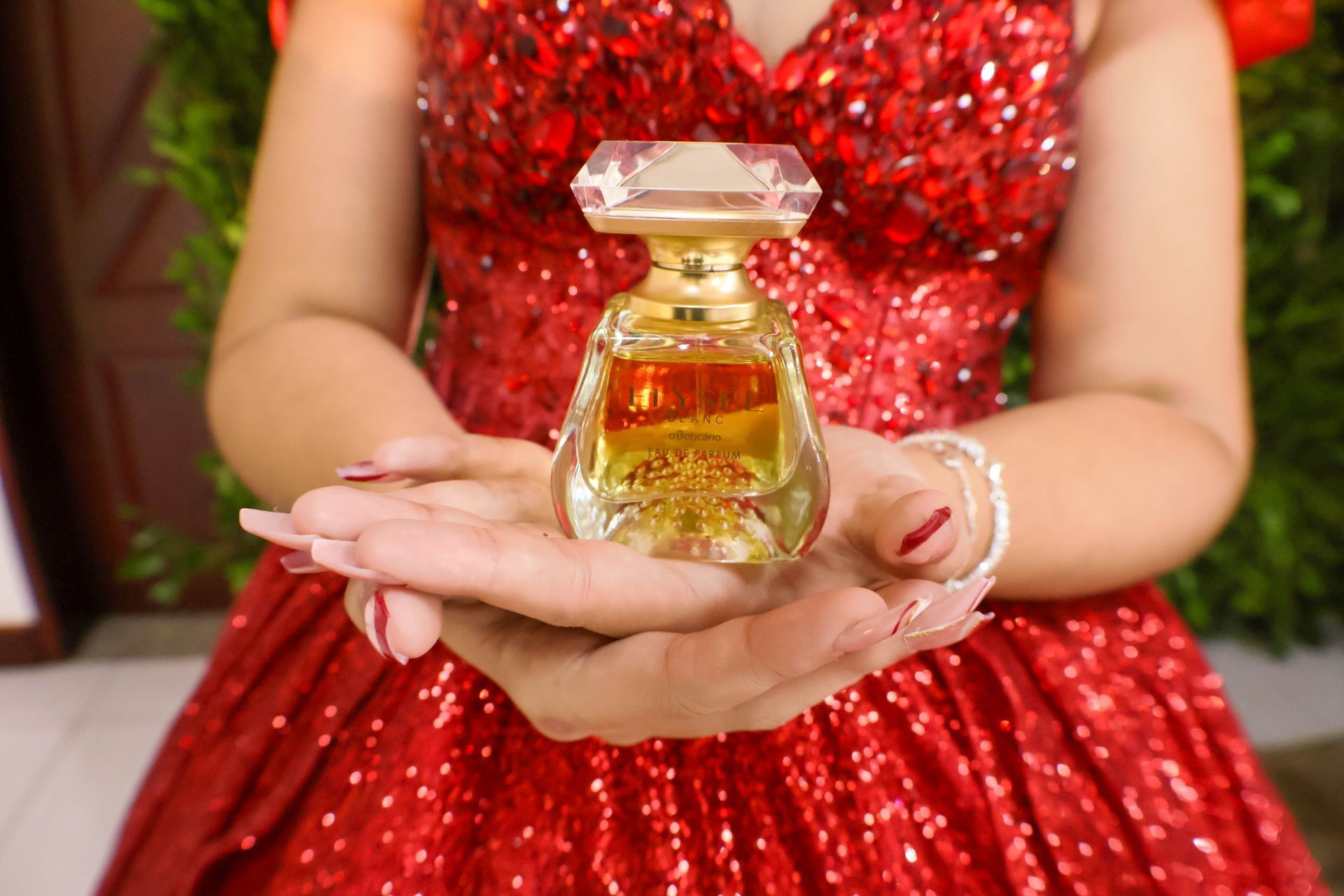 Pessoa com vestido vermelho de lantejoulas segura um frasco de perfume.