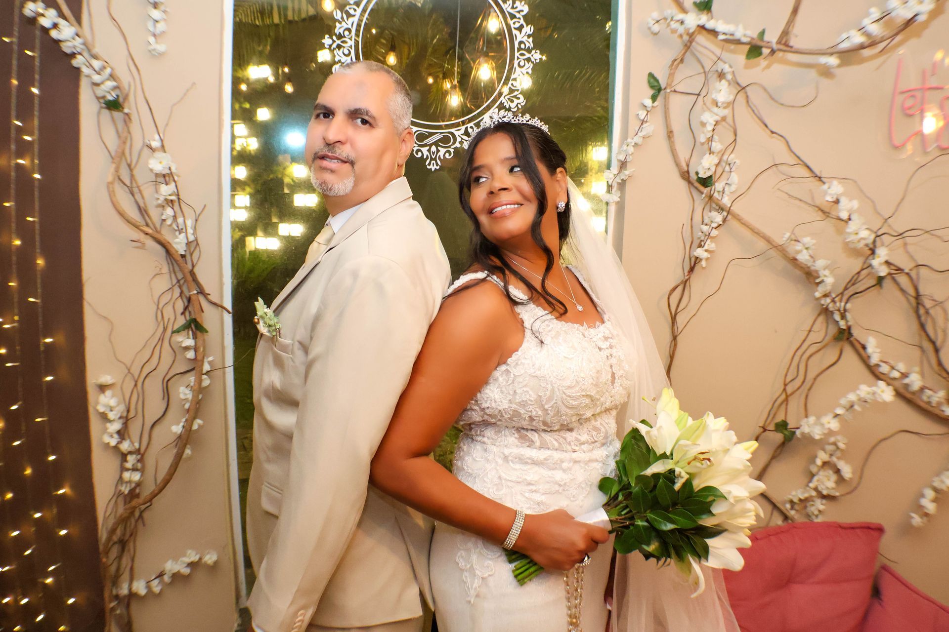 Noivos de costas um para o outro, sorrindo, segurando flores. Traje bege, fundo floral com luzes.