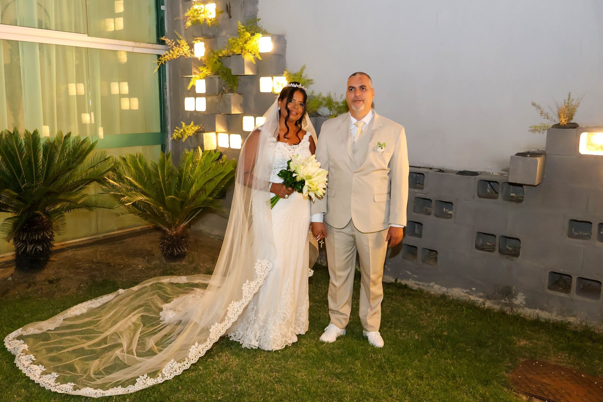 Noivos em trajes de casamento posam ao ar livre, noiva segurando buquê e a mão do noivo sobre a dela. Eles estão na grama.