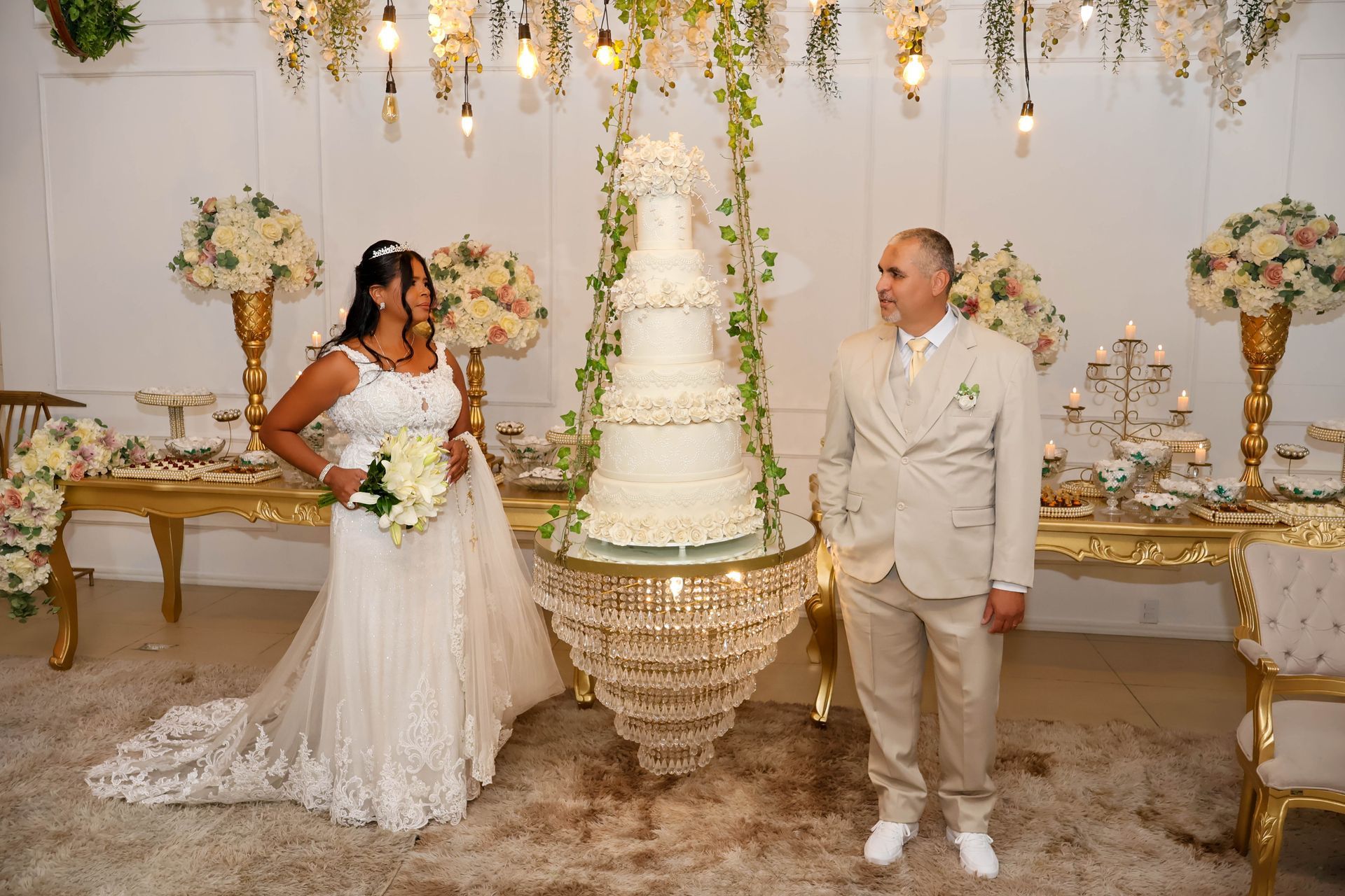 Noivos posam perto de um bolo de casamento de vários andares. Recepção elegante com decoração floral e detalhes dourados.