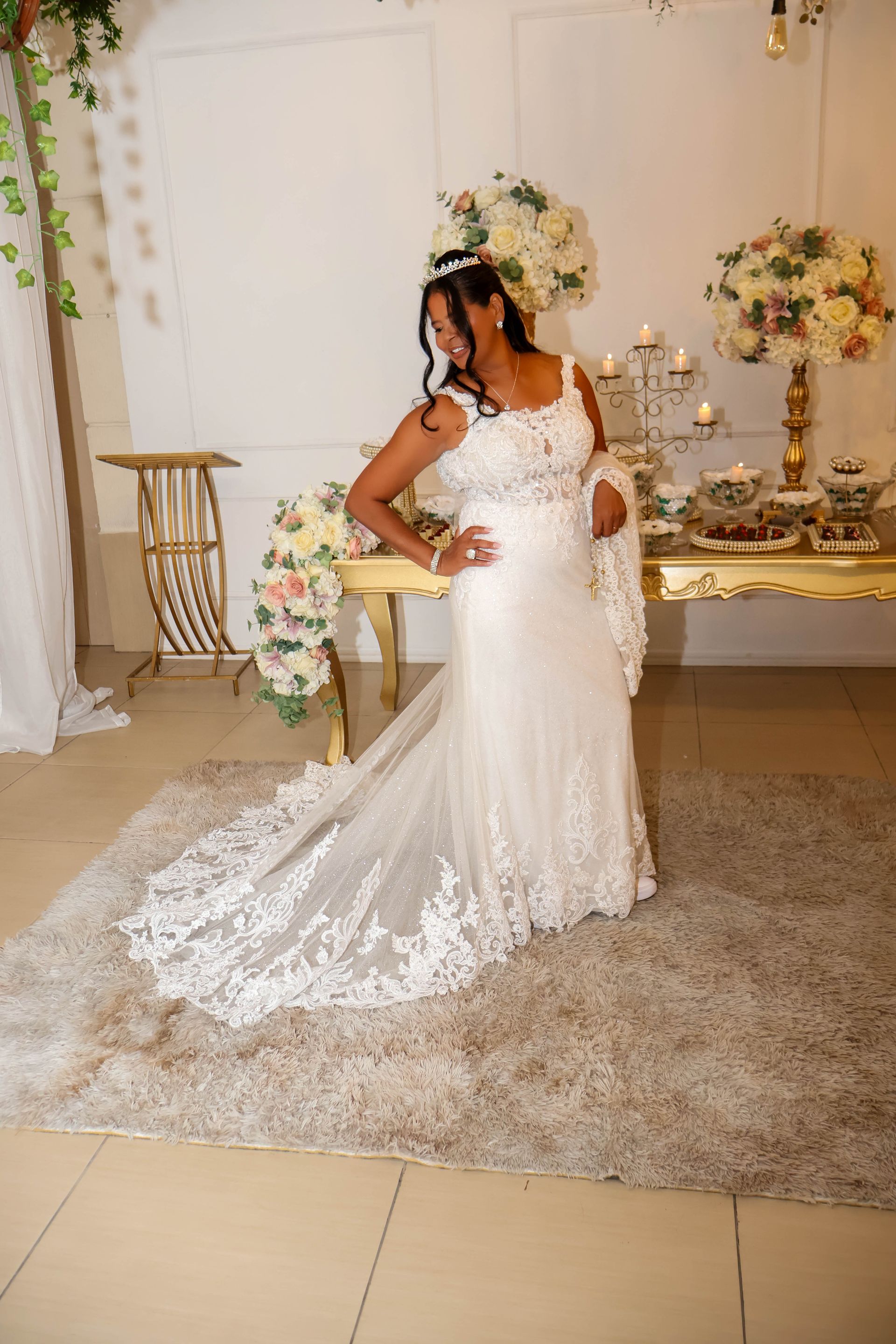 Mulher em um vestido de noiva branco em pé perto de uma mesa com arranjos florais; ambiente interno.