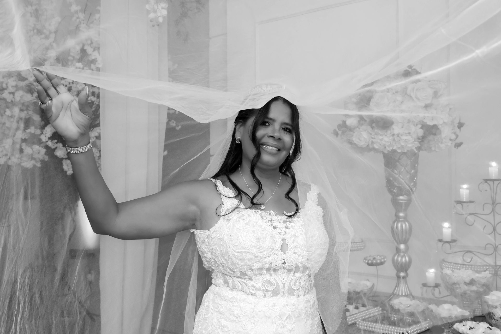 Noiva em vestido de noiva de renda branca, sorrindo, levantando o véu. Ambiente interno com decoração floral.