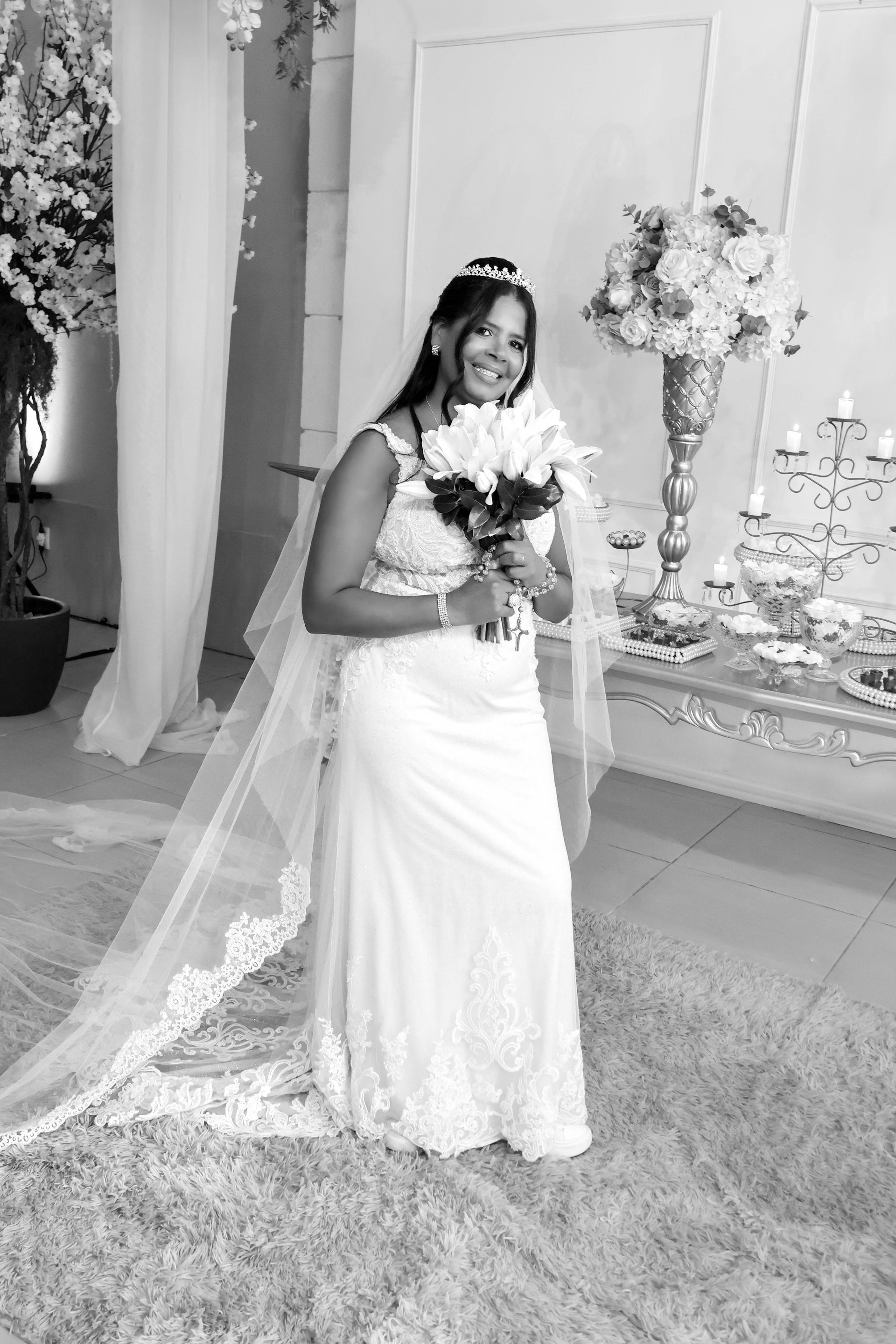 Noiva de vestido branco, segurando flores, sorrindo. Véu longo, decorações florais ao fundo.