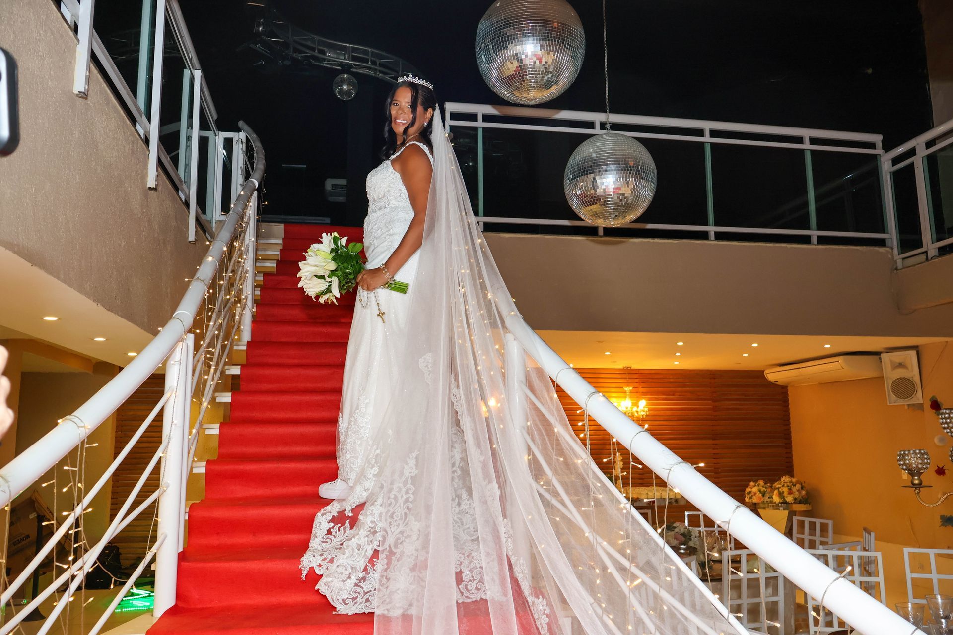 Noiva de vestido branco e véu desce uma escada com carpete vermelho, segurando flores.