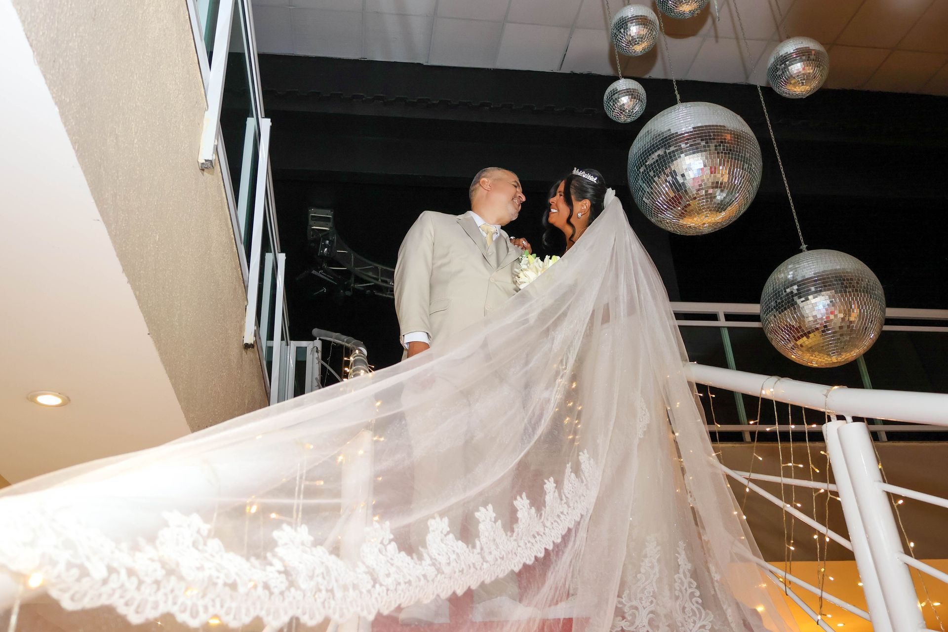 Noivos em pé na escada, sob bolas de discoteca, com o véu da noiva caindo em cascata com detalhes de renda.