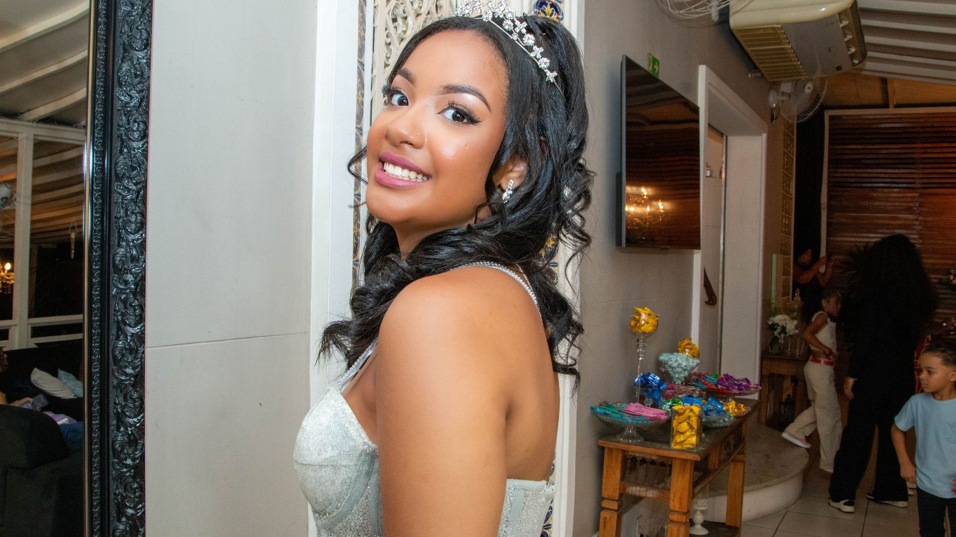 Mulher de cabelos escuros, usando tiara e vestido, sorri, posando em um evento.