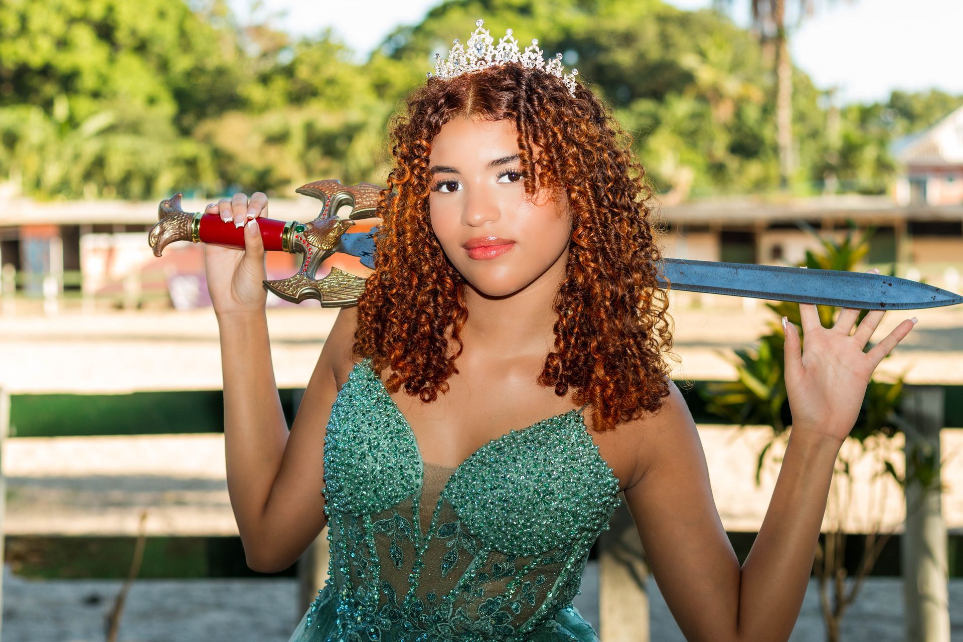 Mulher de vestido verde e tiara segura uma espada, sorrindo. Ao ar livre, luz do sol.