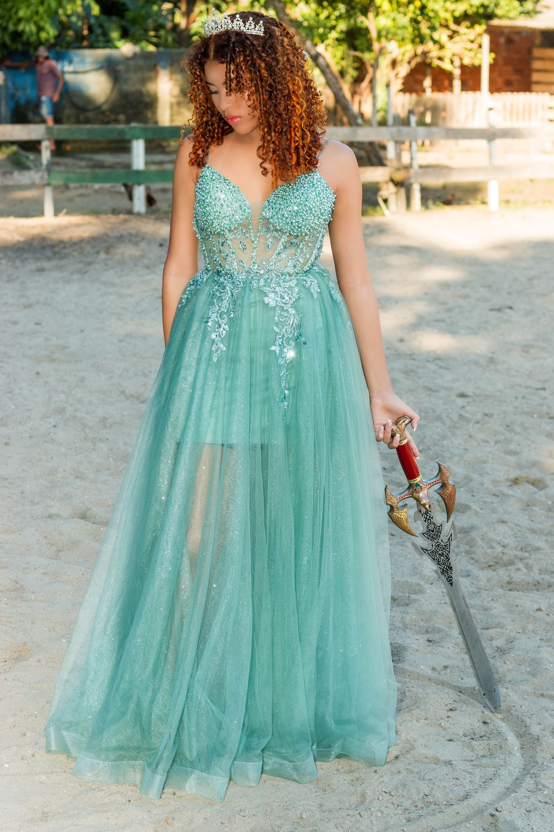 Mulher com um vestido azul-petróleo brilhante e tiara segurando uma espada em uma arena ao ar livre com areia.