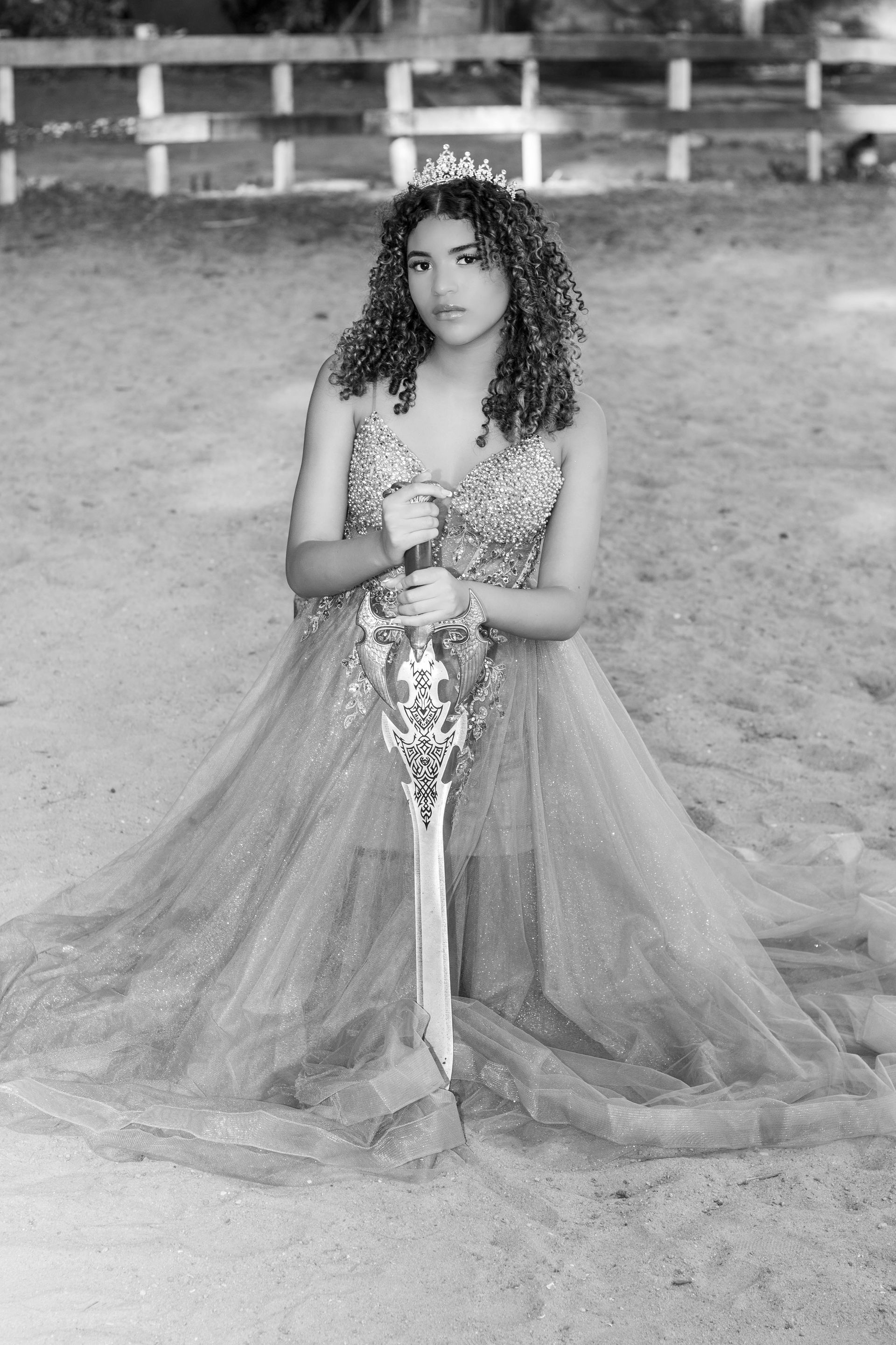 Mulher em traje formal ajoelha-se, segurando uma espada ornamentada. Ela usa uma tiara. O cenário parece ser uma arena ao ar livre.