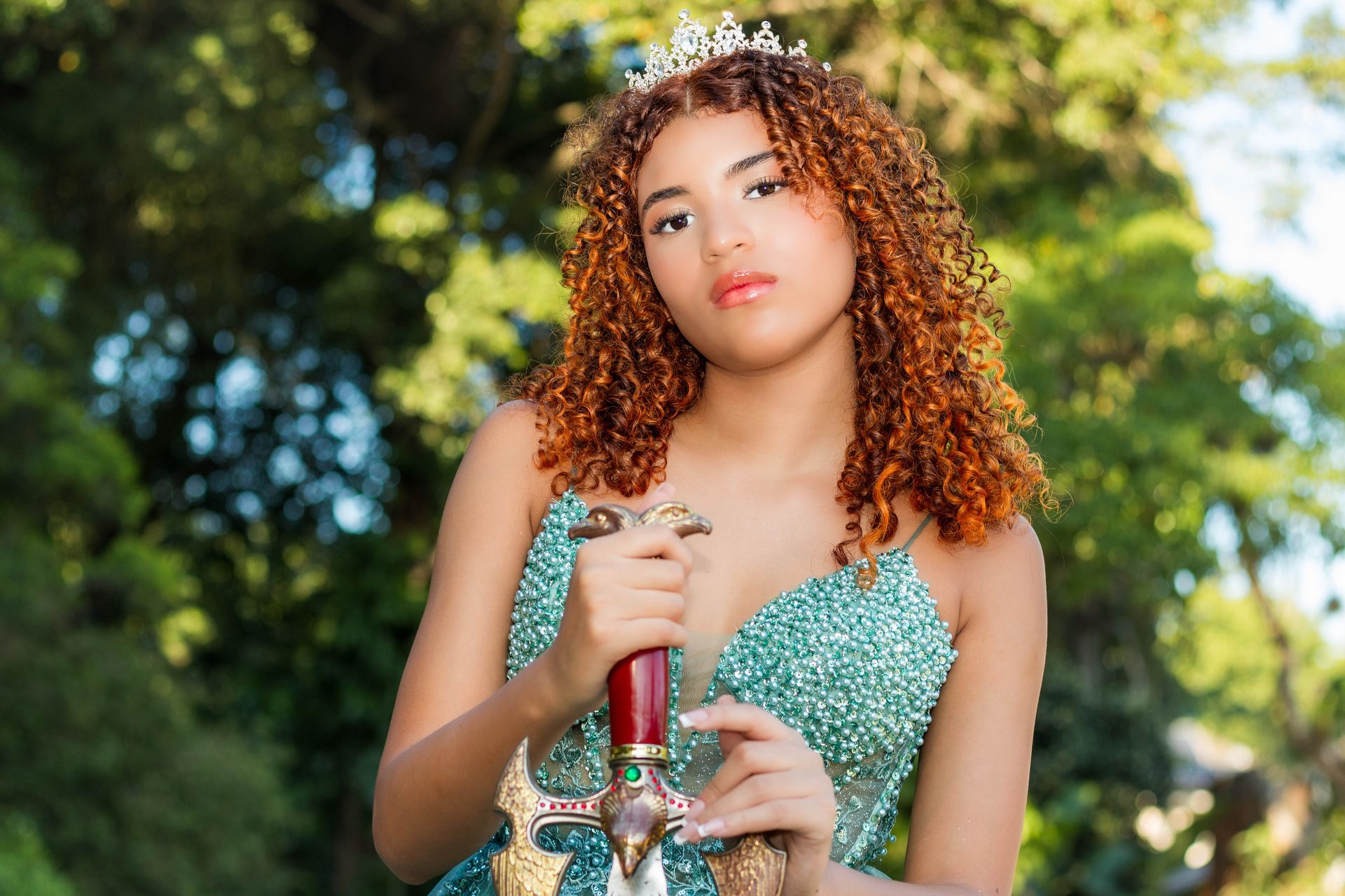 Mulher com vestido azul-petróleo e coroa segura uma espada ao ar livre.