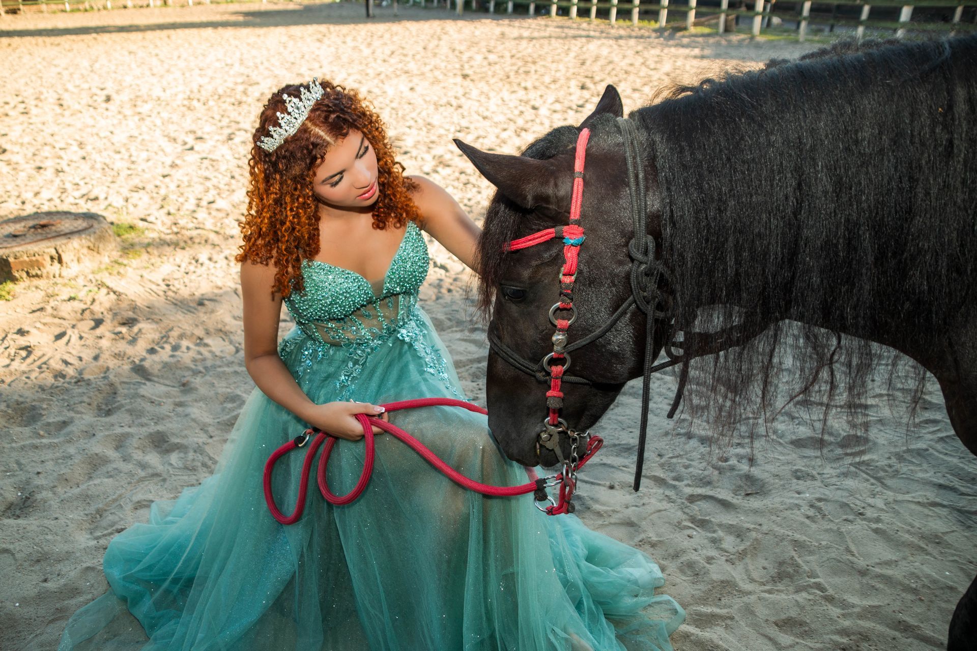 Mulher com vestido azul-petróleo e coroa acaricia um cavalo preto em chão arenoso.