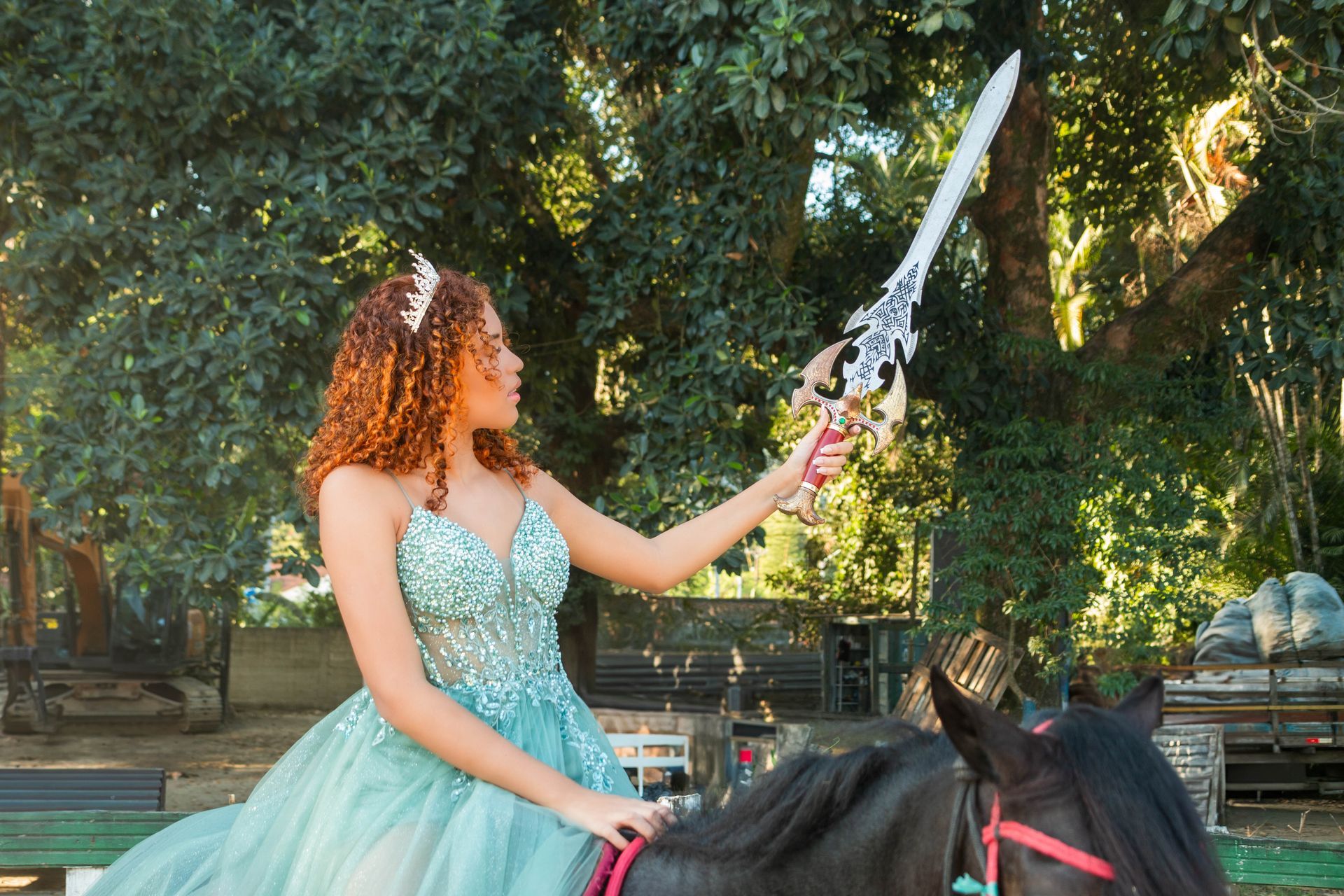 Mulher em vestido azul-petróleo a cavalo ergue uma espada ornamentada, em um ambiente externo com árvores.