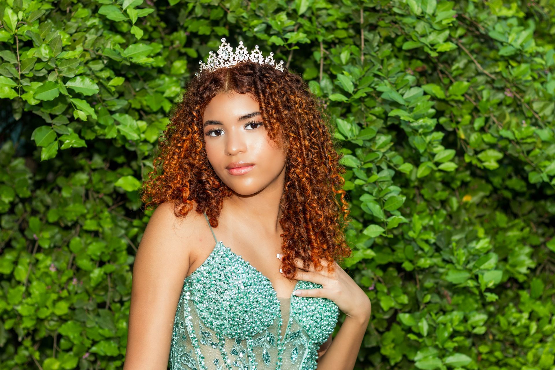 Mulher usando uma tiara e um vestido verde de lantejoulas, posando em frente a uma folhagem verdejante.