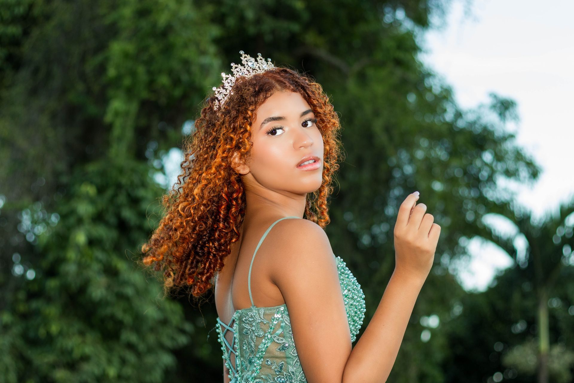 Mulher com cabelos cacheados usando um vestido azul-petróleo e uma coroa, posando ao ar livre.