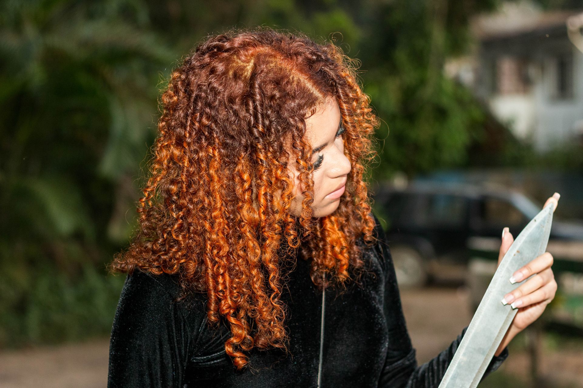 Mulher com cabelo cacheado alaranjado olha para um objeto cinza, ao ar livre.