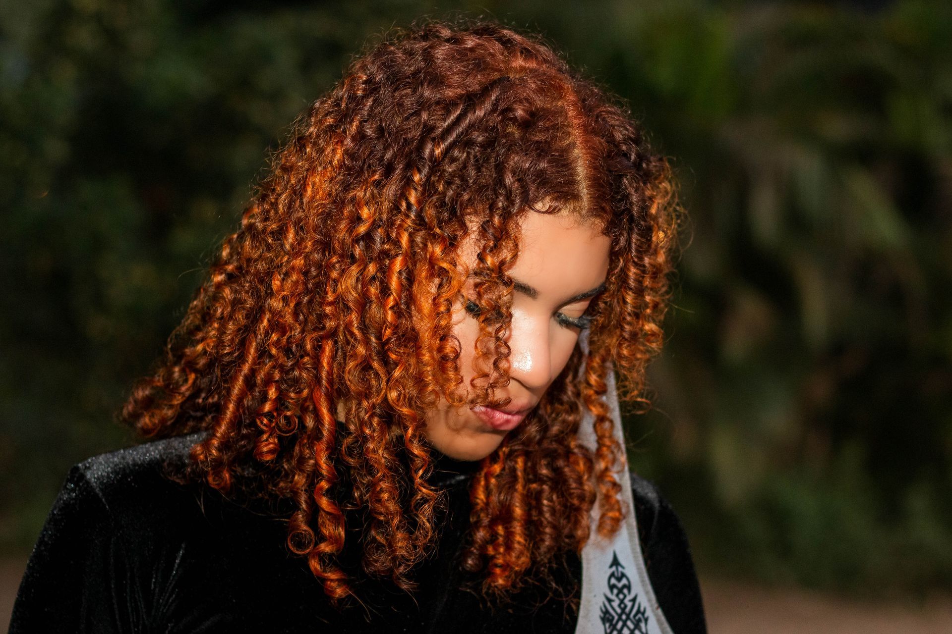 Mulher com cabelos castanho-avermelhados e cacheados, olhando para baixo. Vestindo uma blusa preta contra um fundo desfocado.