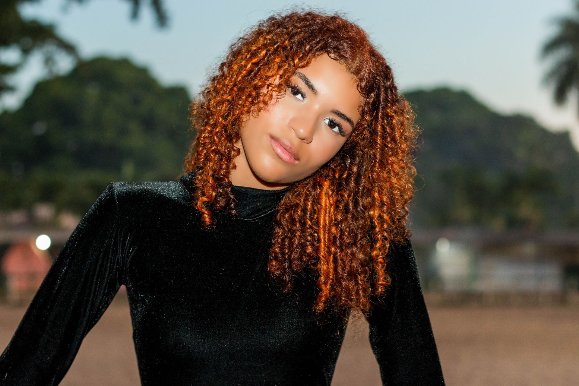 Mulher com cabelo castanho-avermelhado cacheado, vestindo uma blusa preta, ao ar livre com uma expressão neutra.