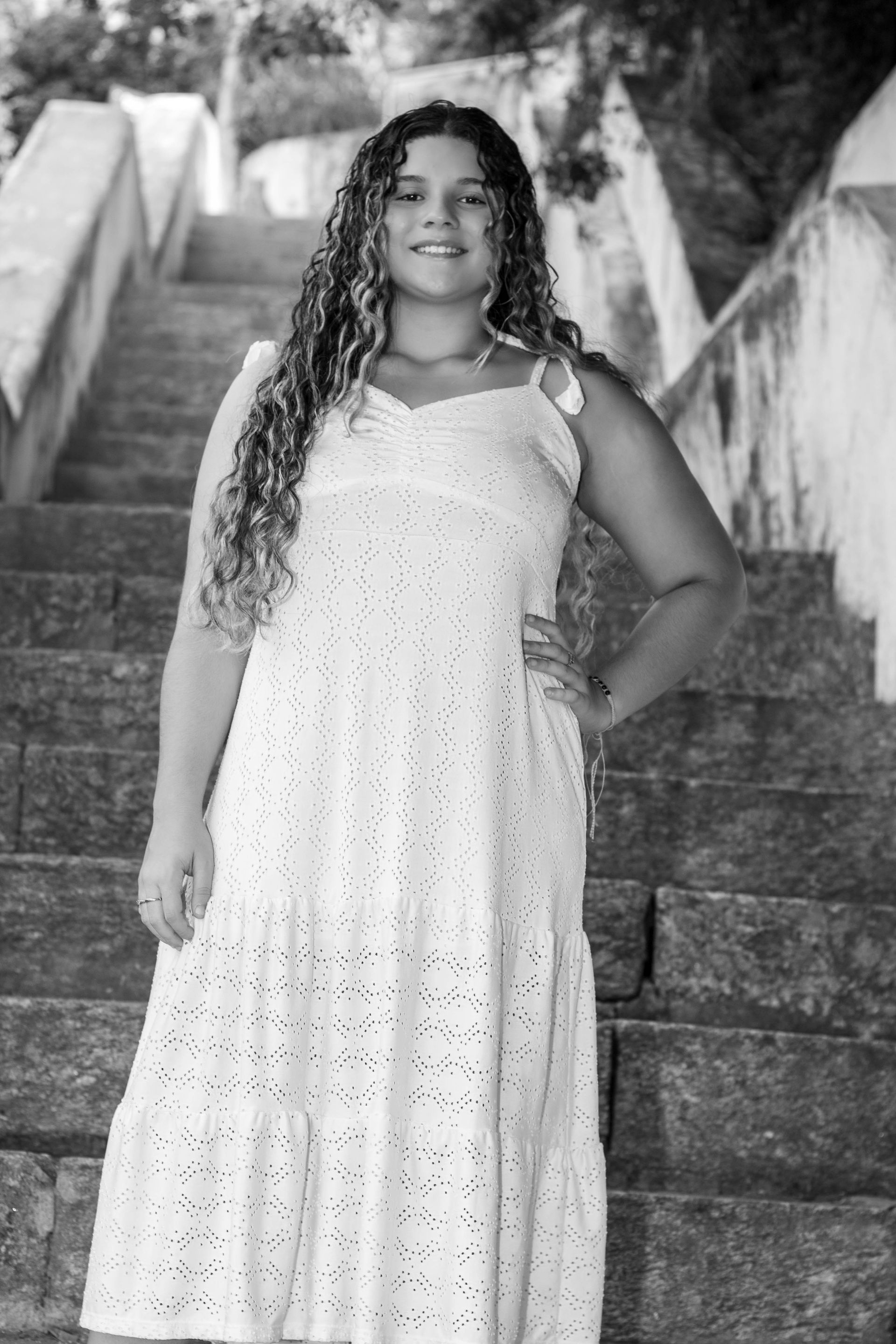Mulher em pé nos degraus, usando um vestido branco, posando com a mão no quadril. Foto em preto e branco.