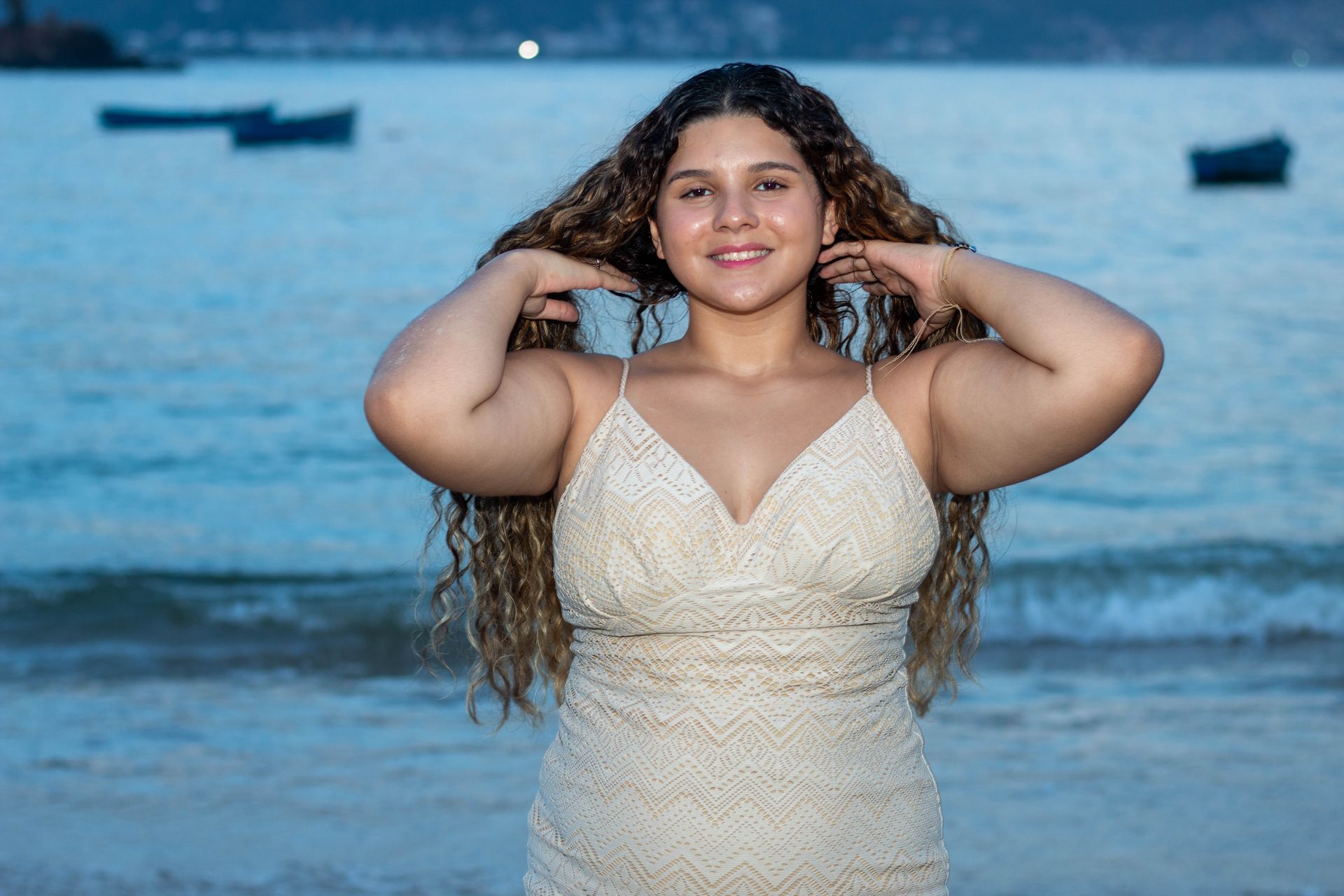 Mulher de cabelos cacheados, de vestido branco, na praia, com as mãos nos cabelos, sorrindo. Oceano e barcos ao fundo.