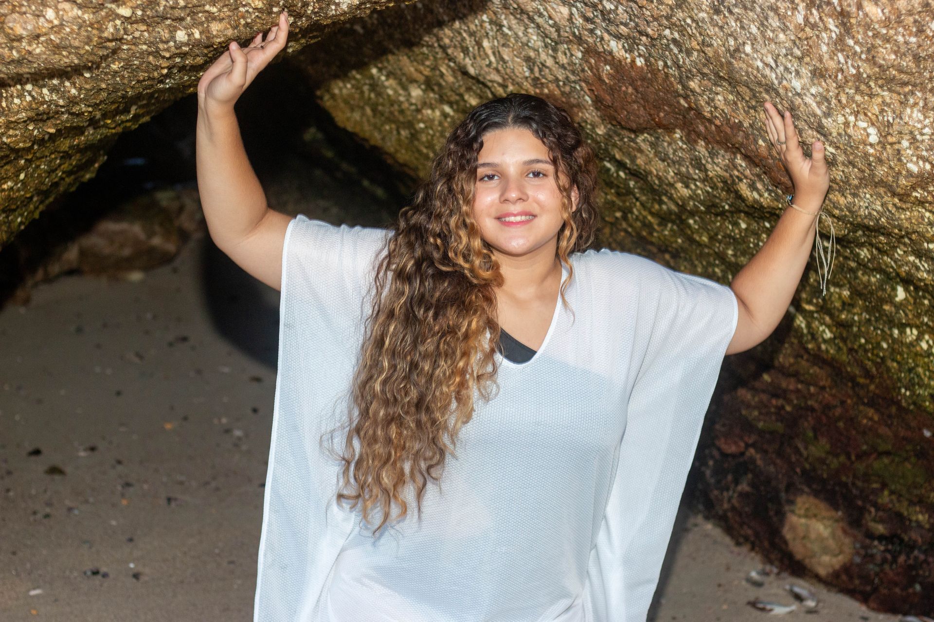Mulher de vestido branco, braços levantados contra uma formação rochosa na praia, sorrindo.