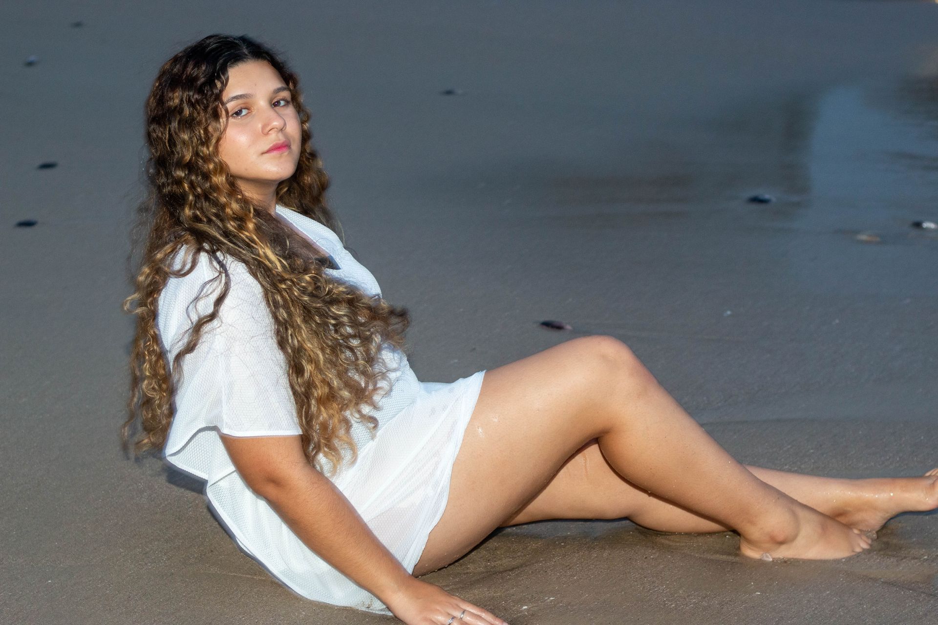 Mulher com cabelos longos e cacheados, de vestido branco, sentada na praia.