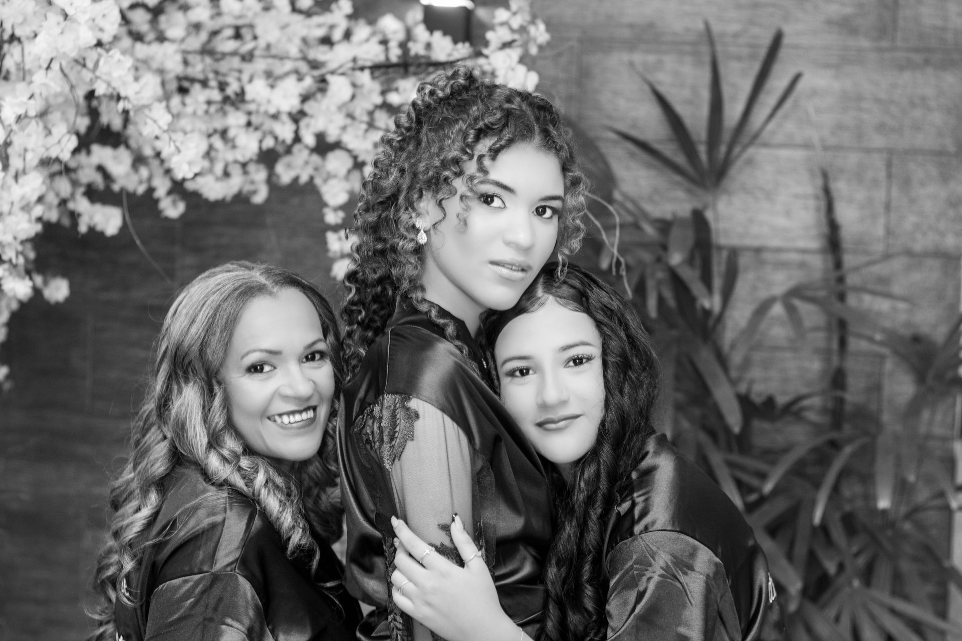 Três pessoas sorrindo e se abraçando. Uma delas tem cabelos cacheados. Elas vestem túnicas e posam perto de plantas e flores.