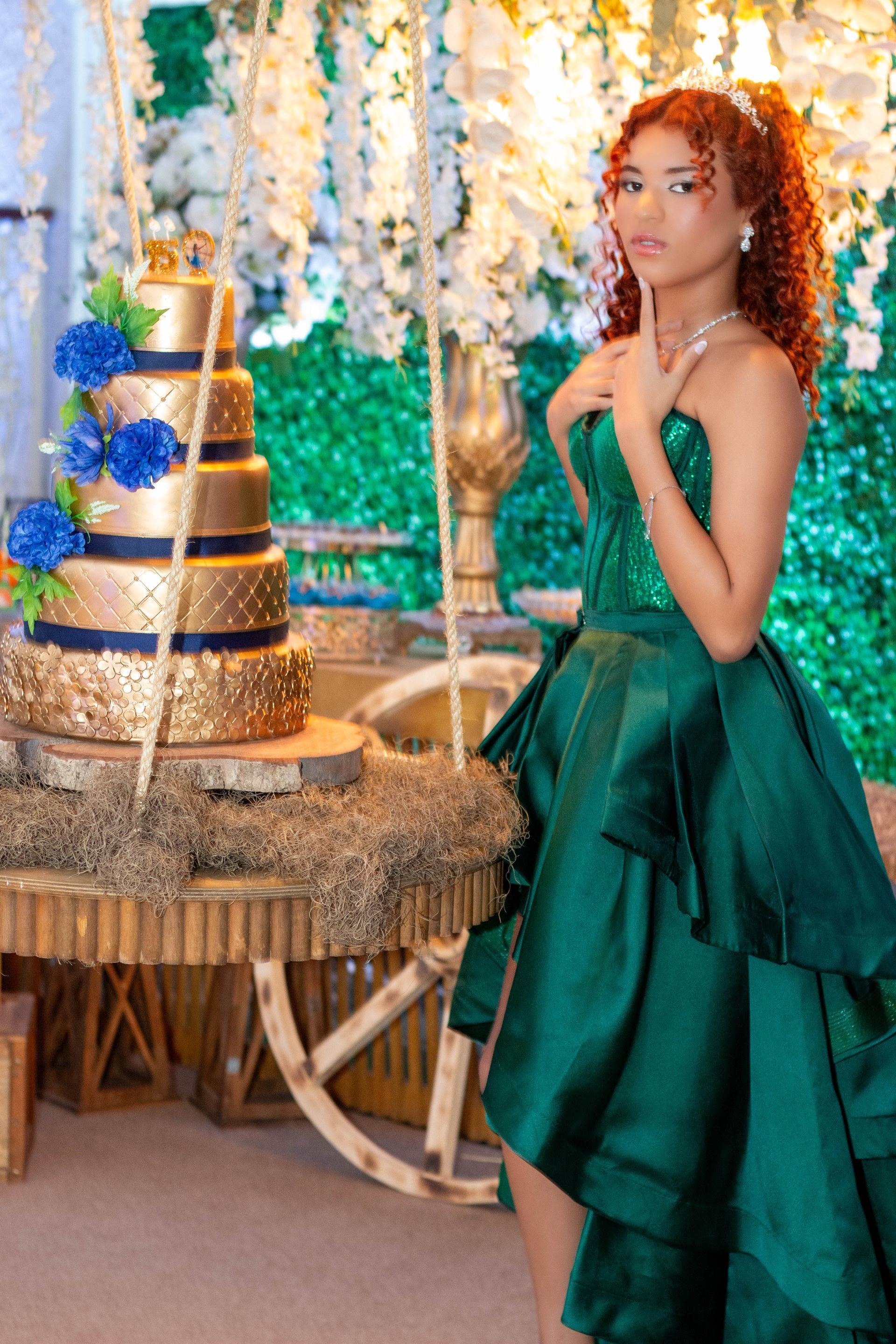 Mulher em vestido esmeralda posa ao lado de um bolo de camadas douradas com flores azuis, em uma festa ao ar livre.