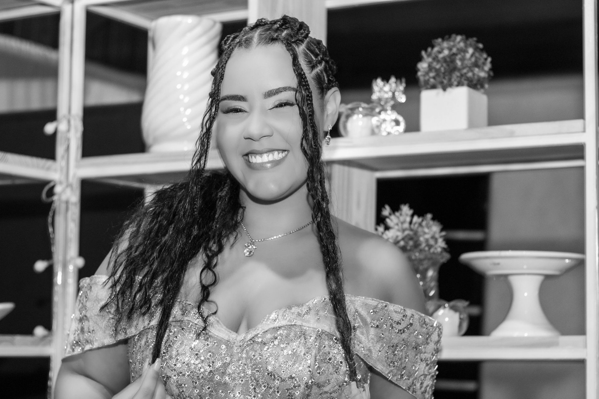 Mulher com cabelo trançado sorrindo, usando um vestido brilhante. Prateleiras com decorações ao fundo.